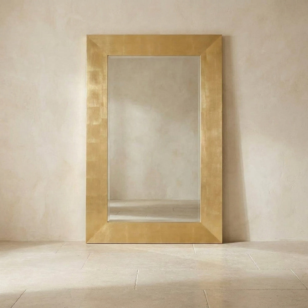 A large rectangular wall mirror with a wide, textured metallic gold frame, leaning against a neutral beige plaster wall on a light stone floor.