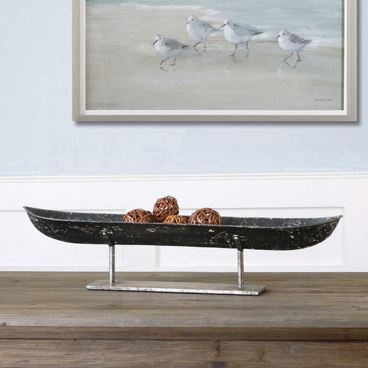 Long black bowl for decoration on a rustic wood console with framed coastal art hung above.