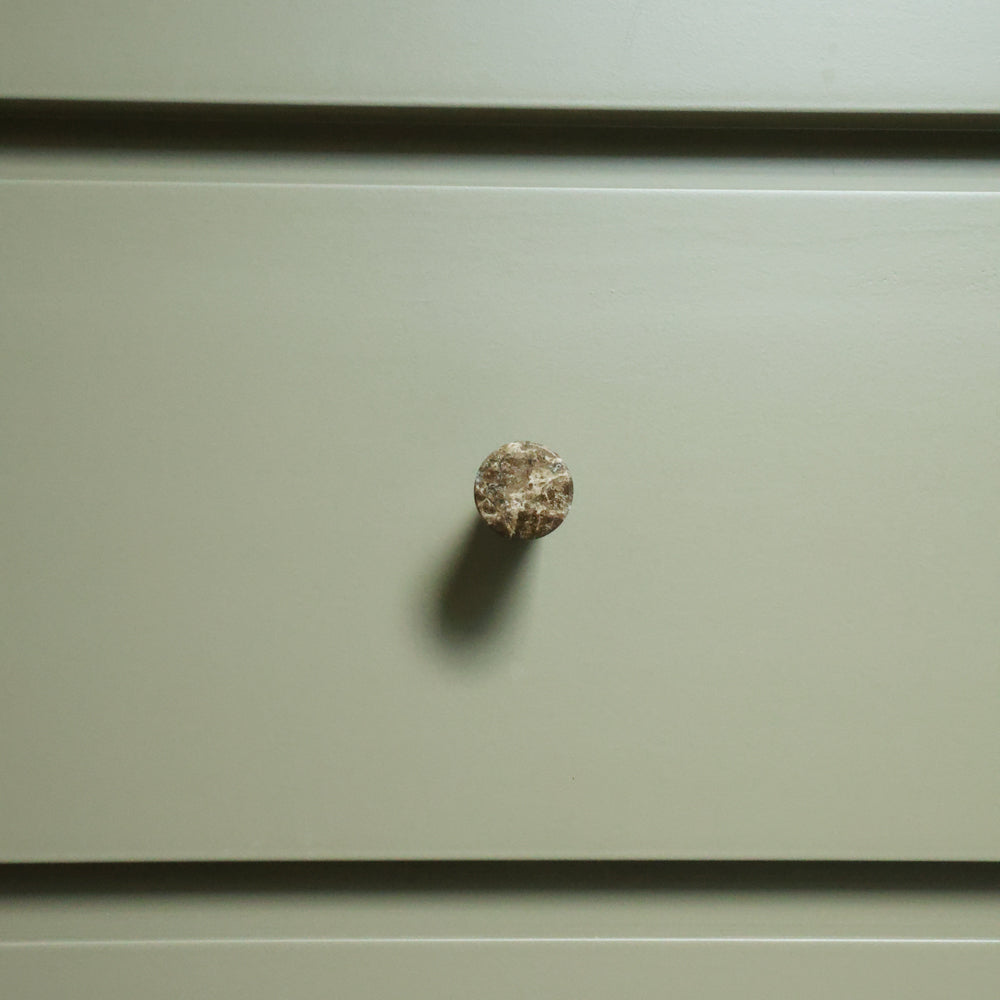 A small round, textured marble knob mounted on a flat, muted sage green drawer front.
