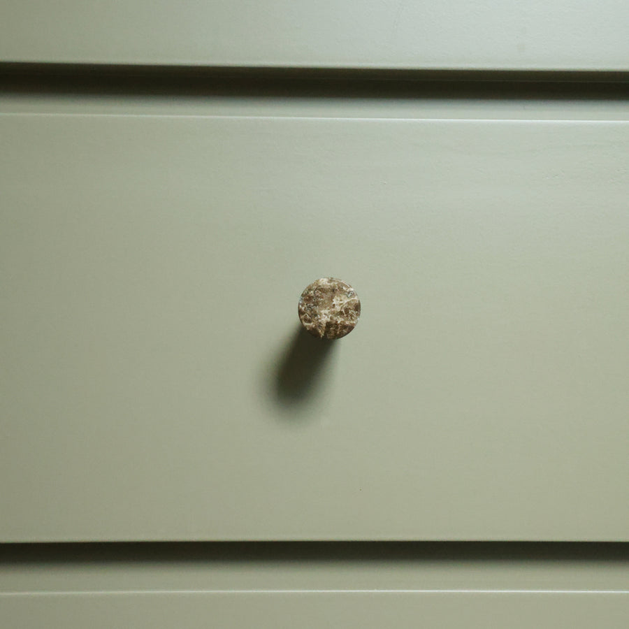 A small round, textured marble knob mounted on a flat, muted sage green drawer front.