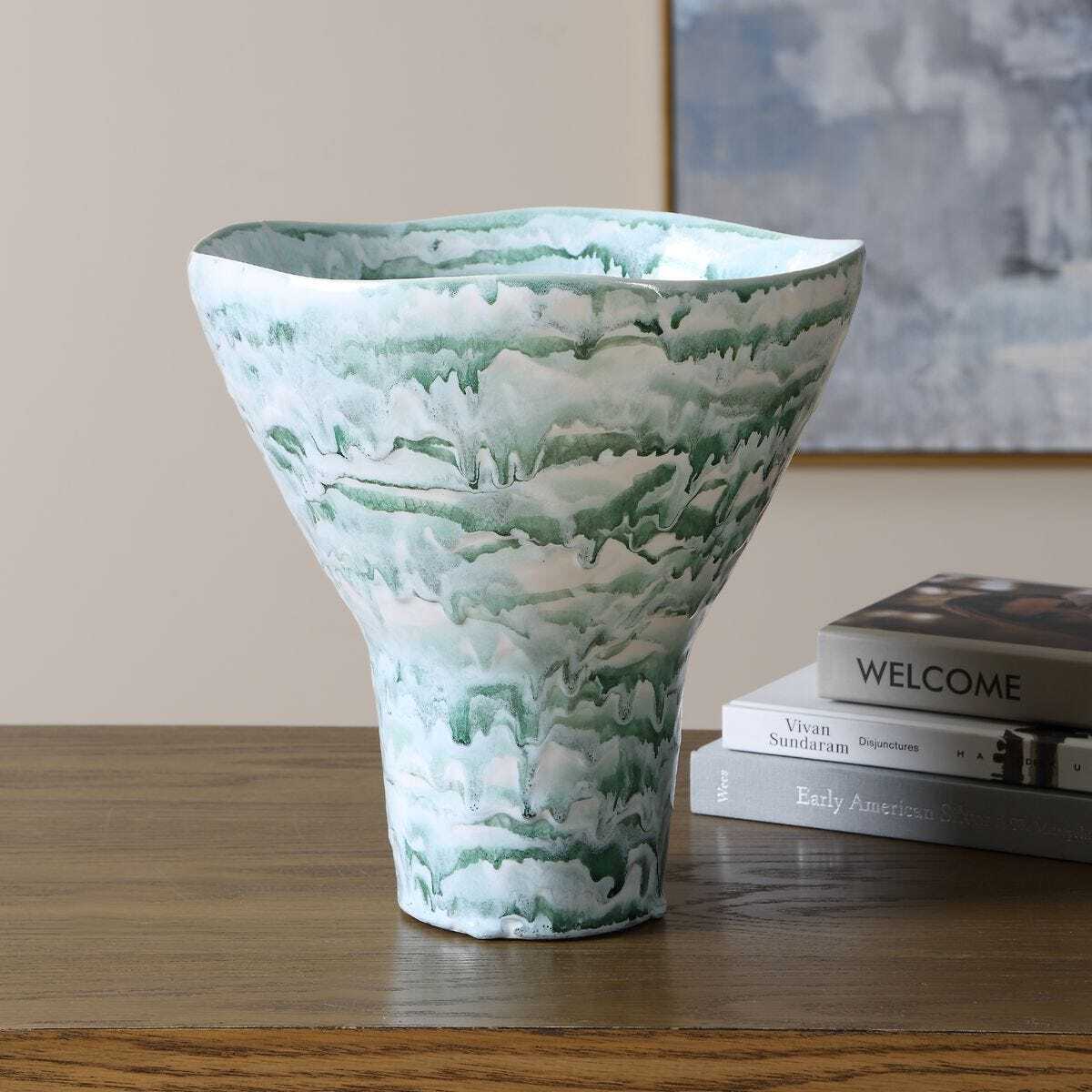 A sculptural ceramic vase with a green and white marbled pattern, displayed on a wooden table next to a stack of books in an indoor setting.