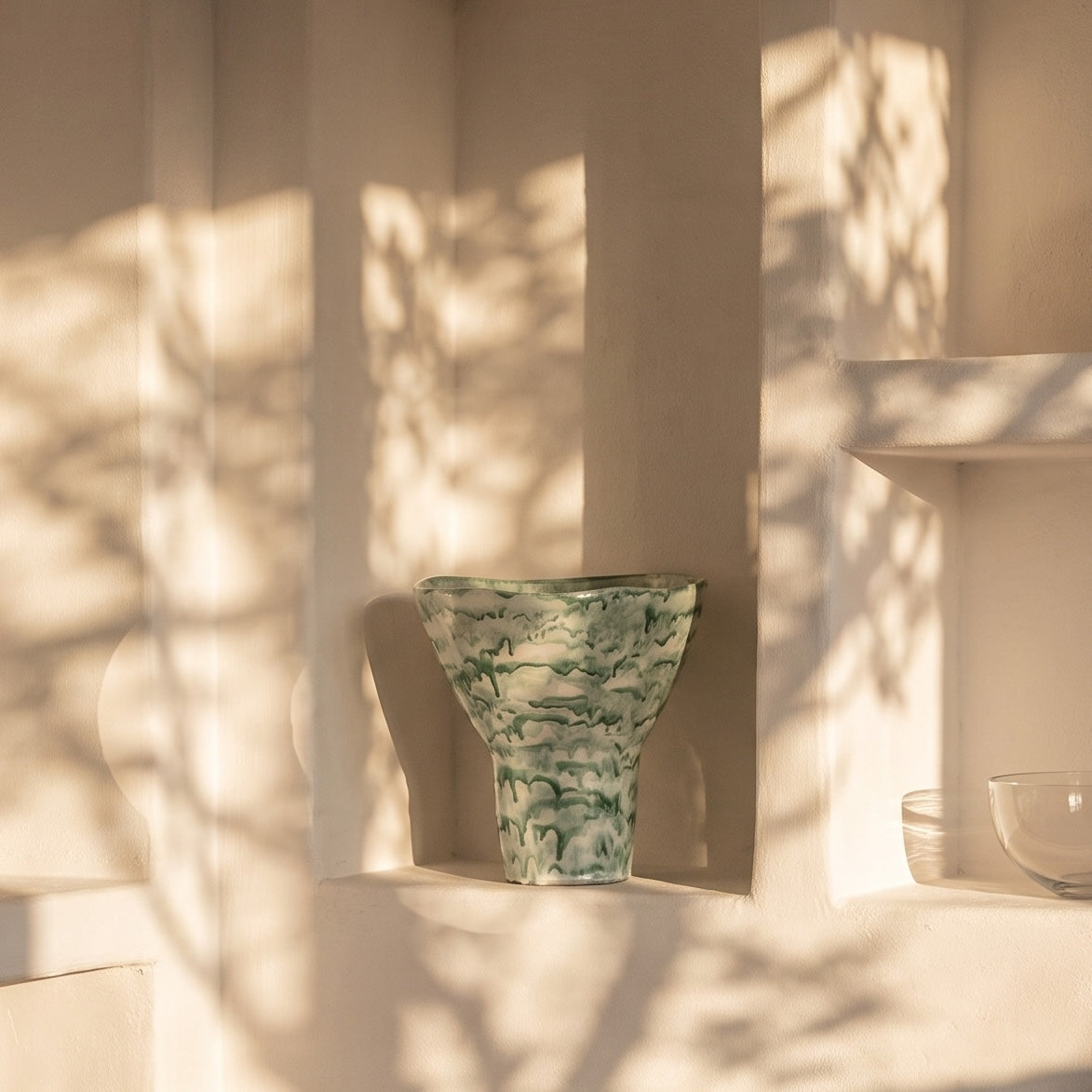 A green and white marbled ceramic vase displayed on a cream-colored shelf, with dappled sunlight and leaf shadows cast across the background.