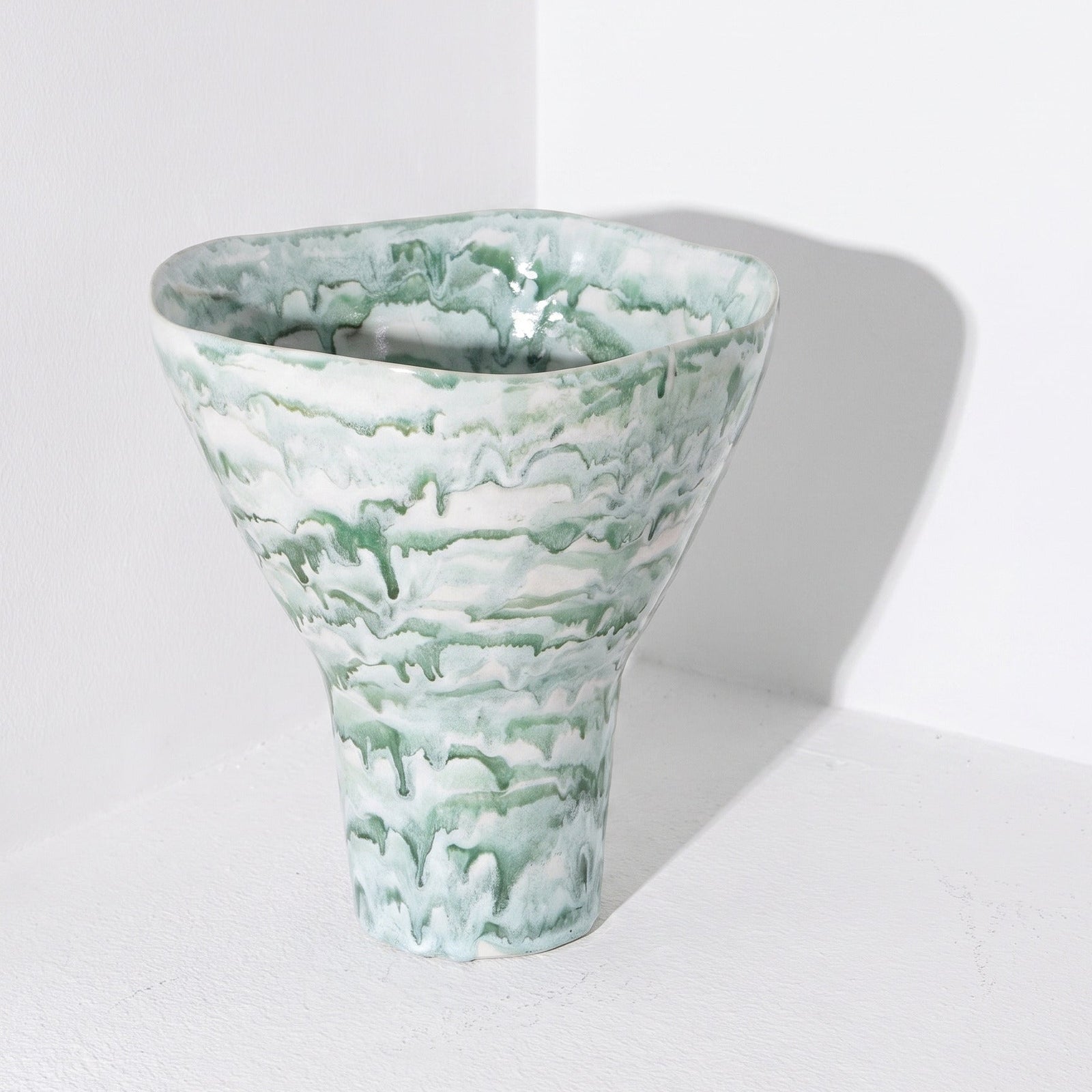 A sculptural ceramic vase with a wide, flared top and a narrow pedestal base, featuring a marbled, cloud-like pattern in sage green and white. It is photographed in a minimalist white corner studio setting with a soft shadow cast to the right.
