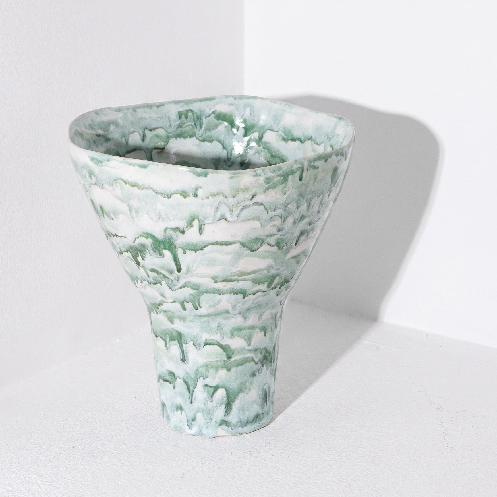 A sculptural ceramic vase with a wide, flared top and a narrow pedestal base, featuring a marbled, cloud-like pattern in sage green and white. It is photographed in a minimalist white corner studio setting with a soft shadow cast to the right.