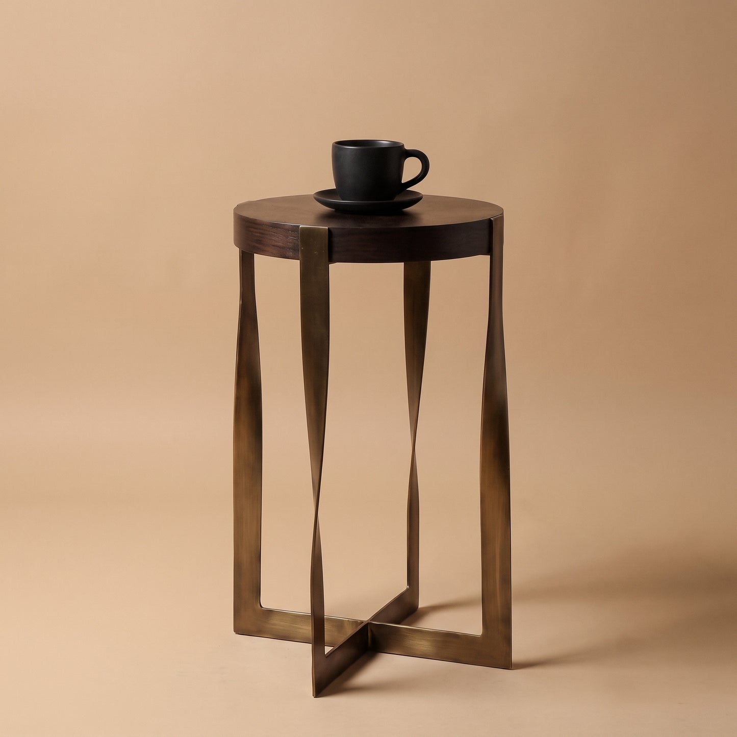 A small round side table with a dark wood top and twisted metal legs in a bronze finish, shown on a warm beige studio background with a black cup and saucer placed on top.