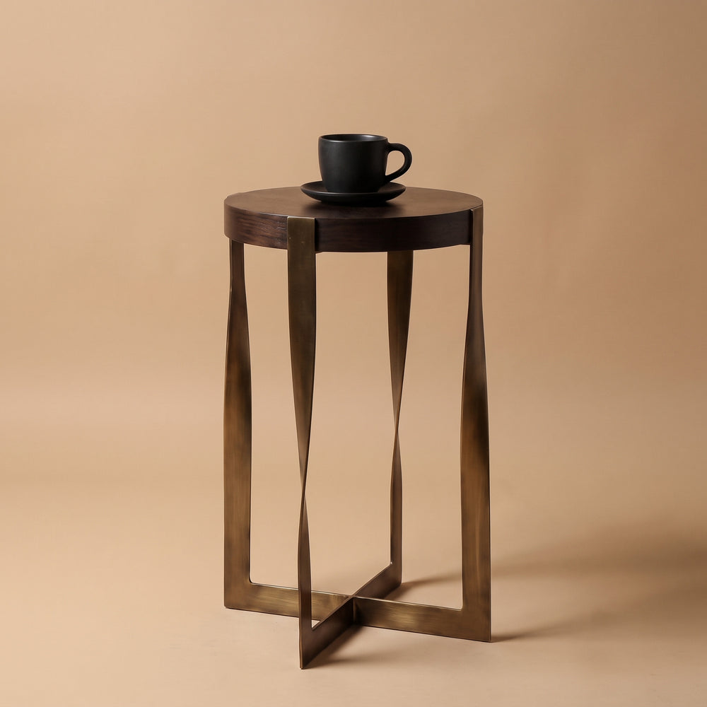 A small round side table with a dark wood top and twisted metal legs in a bronze finish, shown on a warm beige studio background with a black cup and saucer placed on top.