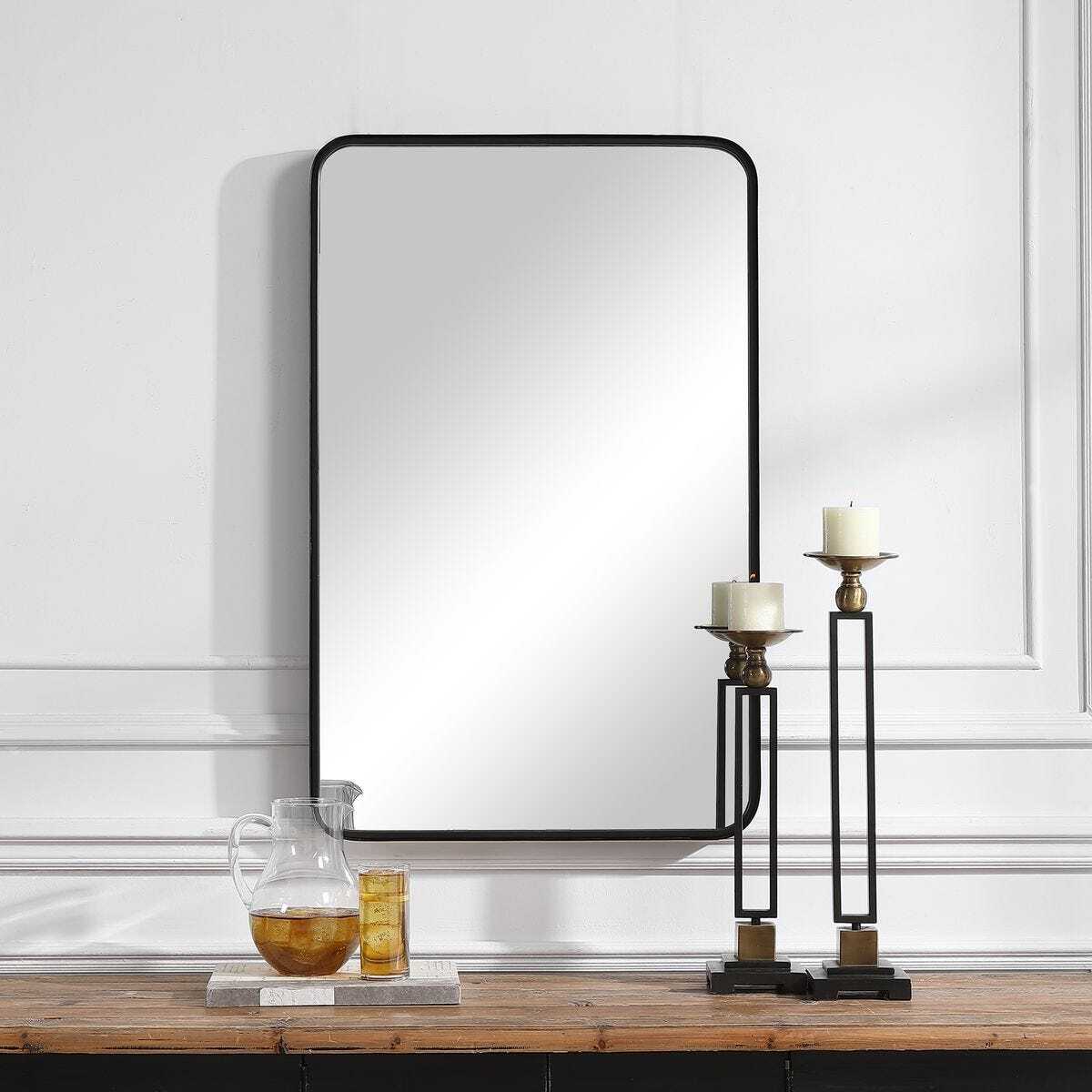 Rectangular mirror with black frame on a white wall above a wooden surface with decorative items.