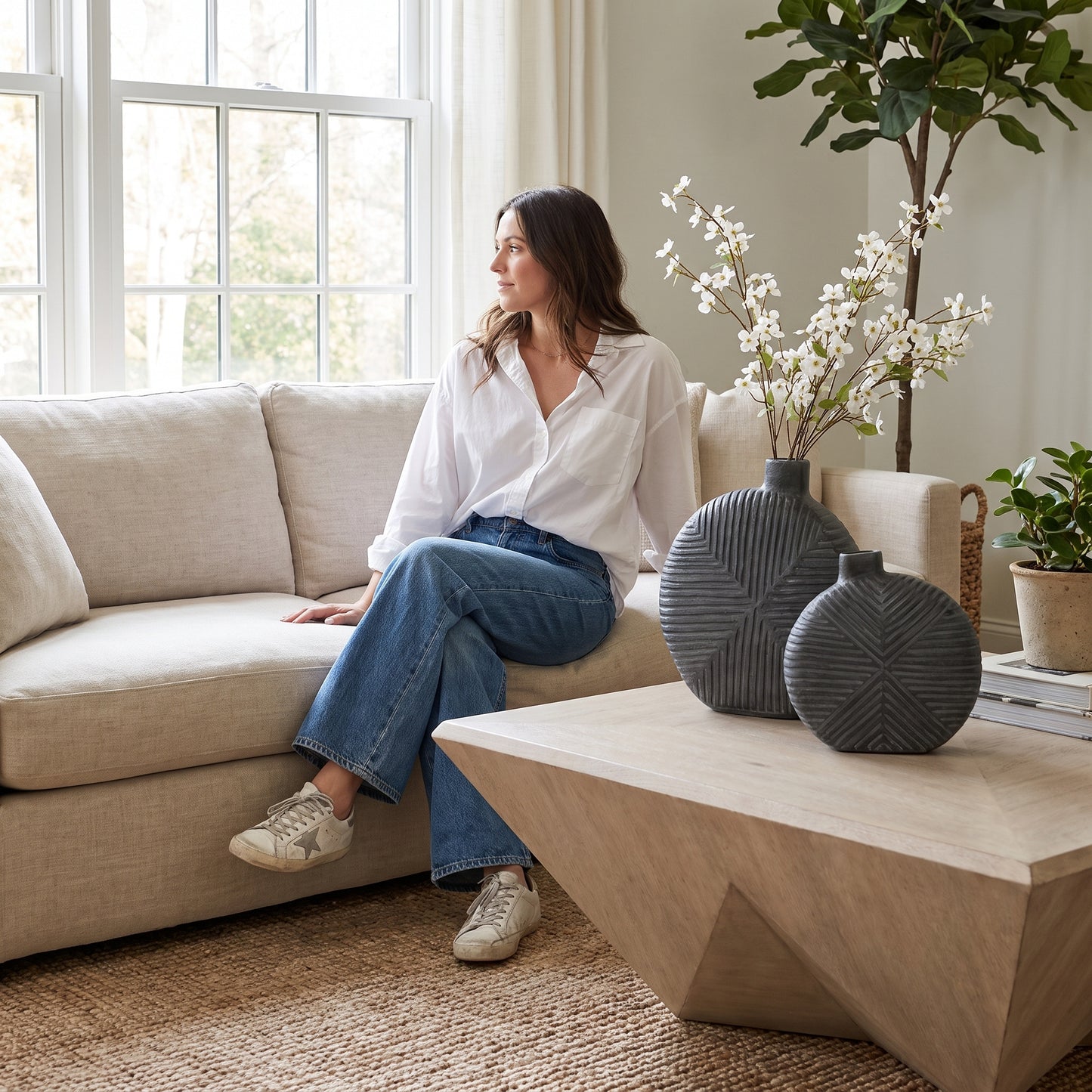 A woman sits on a light beige sofa in a bright living room, wearing a white button-up shirt and blue jeans. In the foreground, a light wood coffee table holds two dark gray ribbed decorative vases with white blossoms in the background.