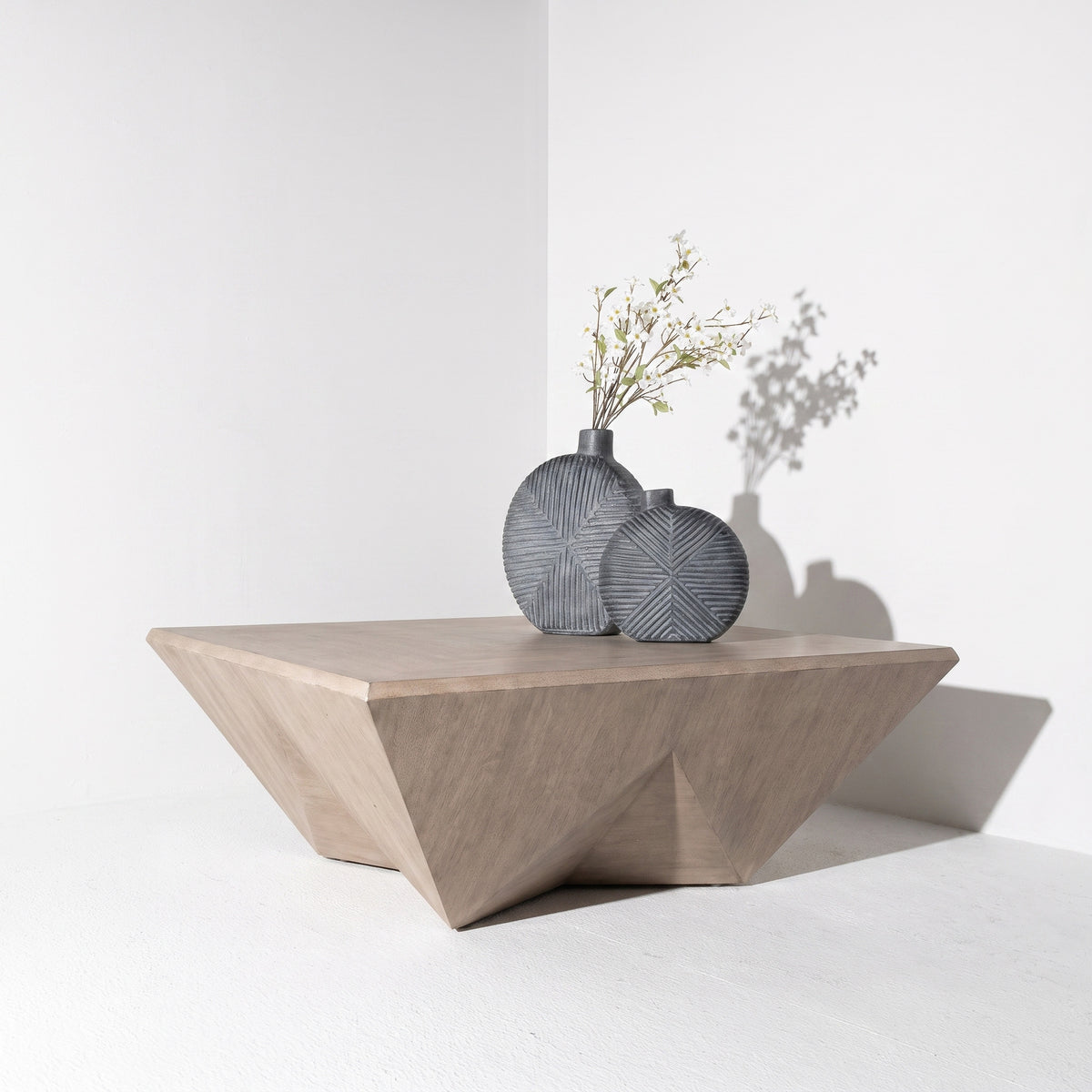 A modern geometric wooden side table in a light oak finish, styled with two textured gray ceramic vases containing dried branches, set against a clean white wall with soft shadows.