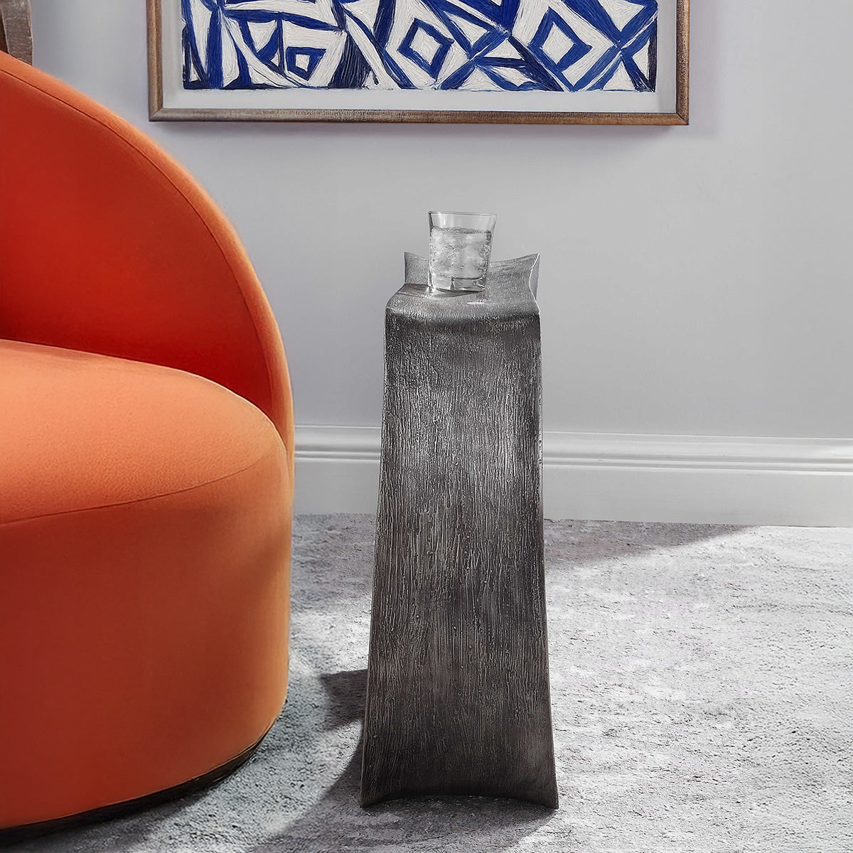 Modern gray accent able with water on top next to an orange chair in a living room with blue abstract art.