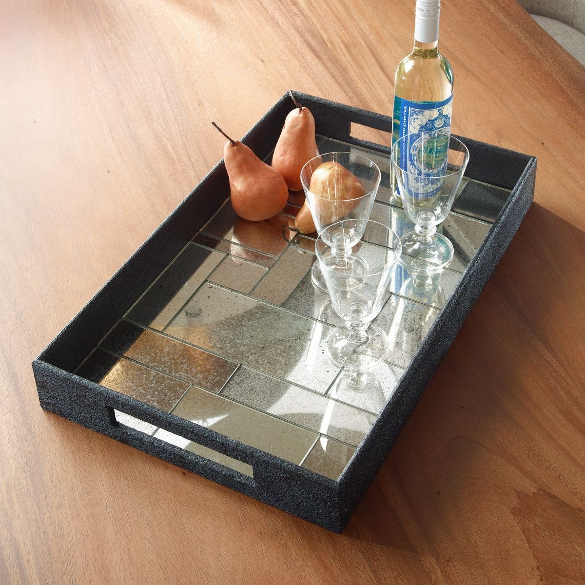 Decorative tray with reflective surface holding a bottle, glasses, and pears on a wooden surface.
