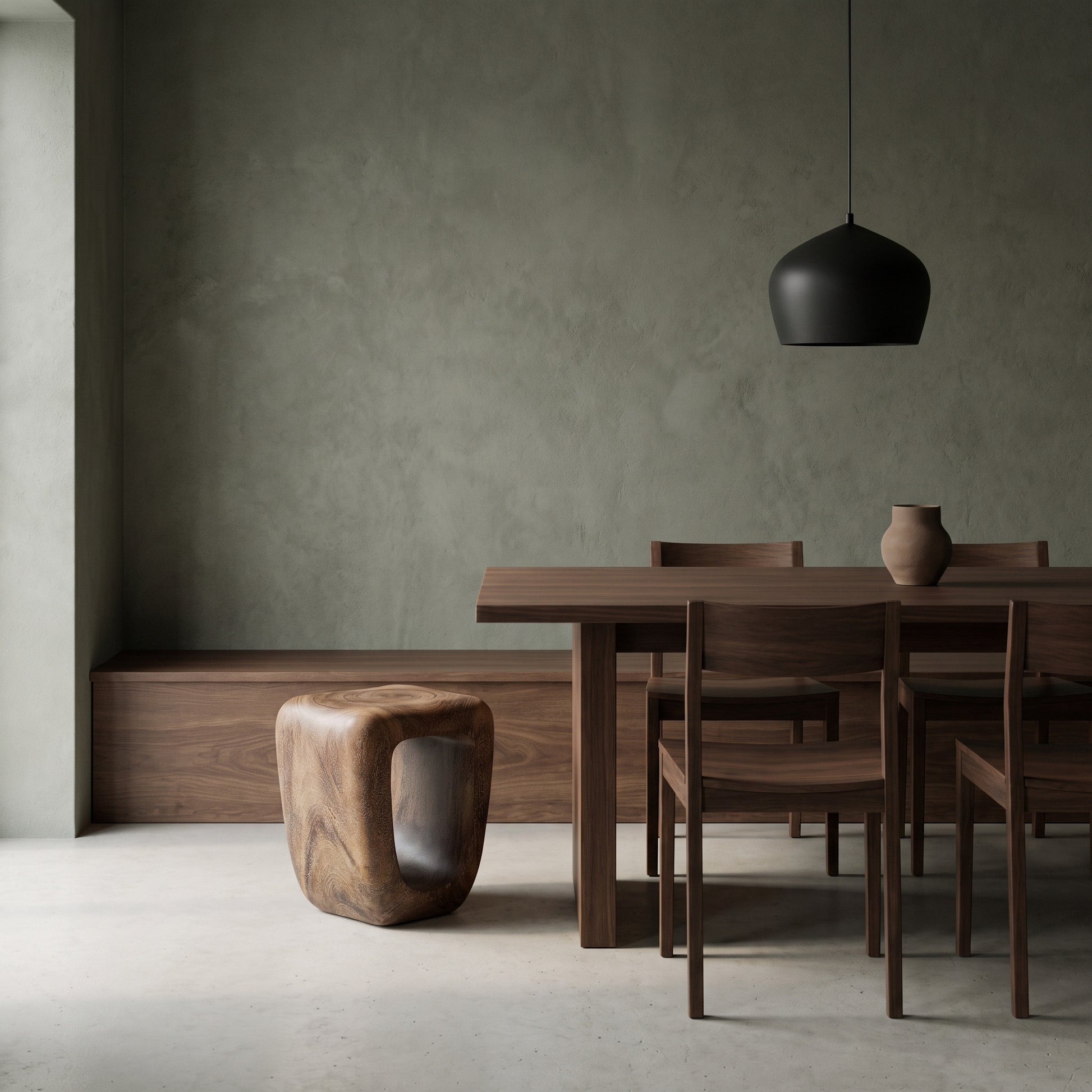 A modern dining room scene featuring a dark wood dining table and chairs, a sculptural wooden stool, a black pendant light, and a textured gray wall.