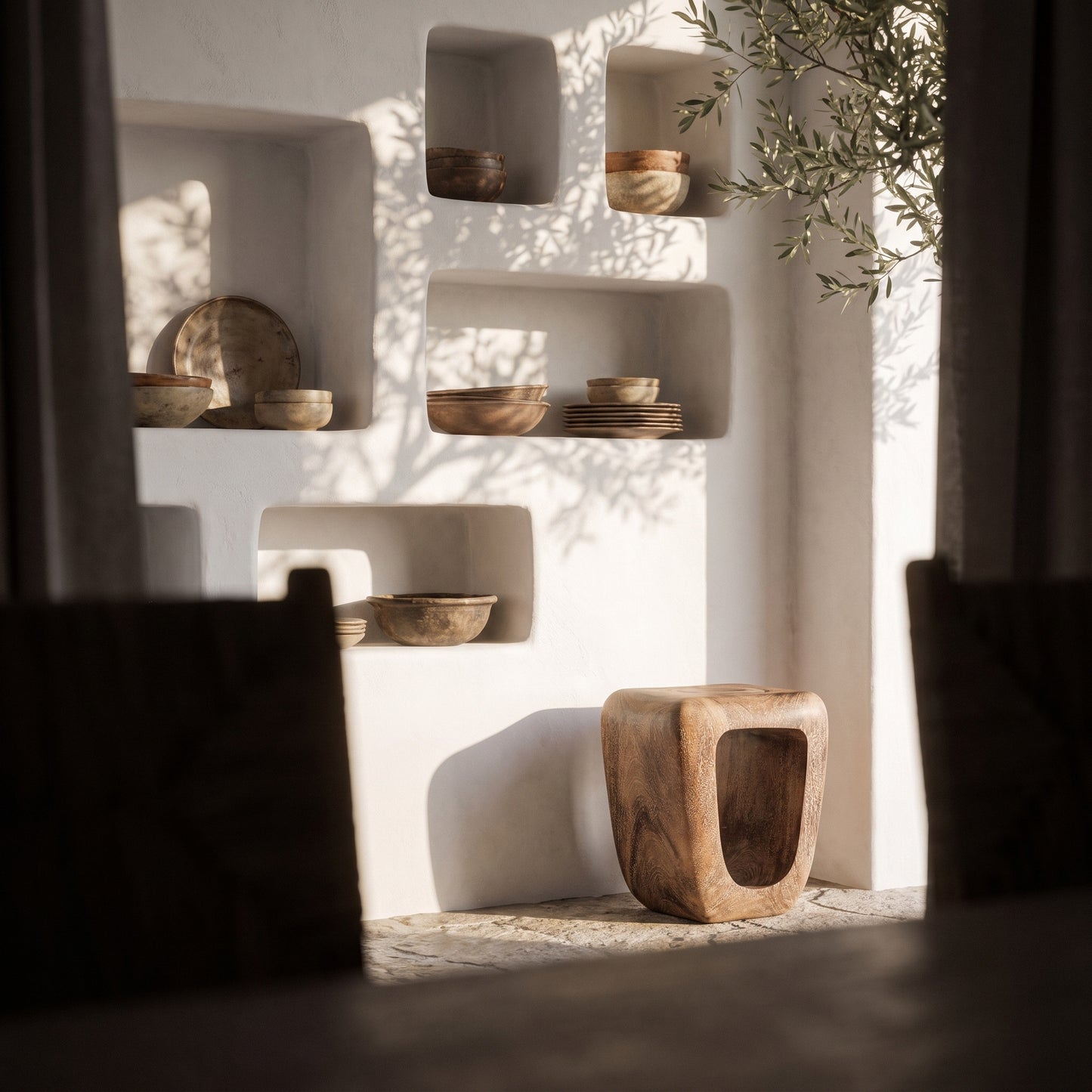 A rustic wooden stool with a hollow center sits on a stone floor in front of a white stucco wall with recessed shelves holding bowls and plates. Sunlight casts leafy shadows across the wall, with dark curtains framing the scene.