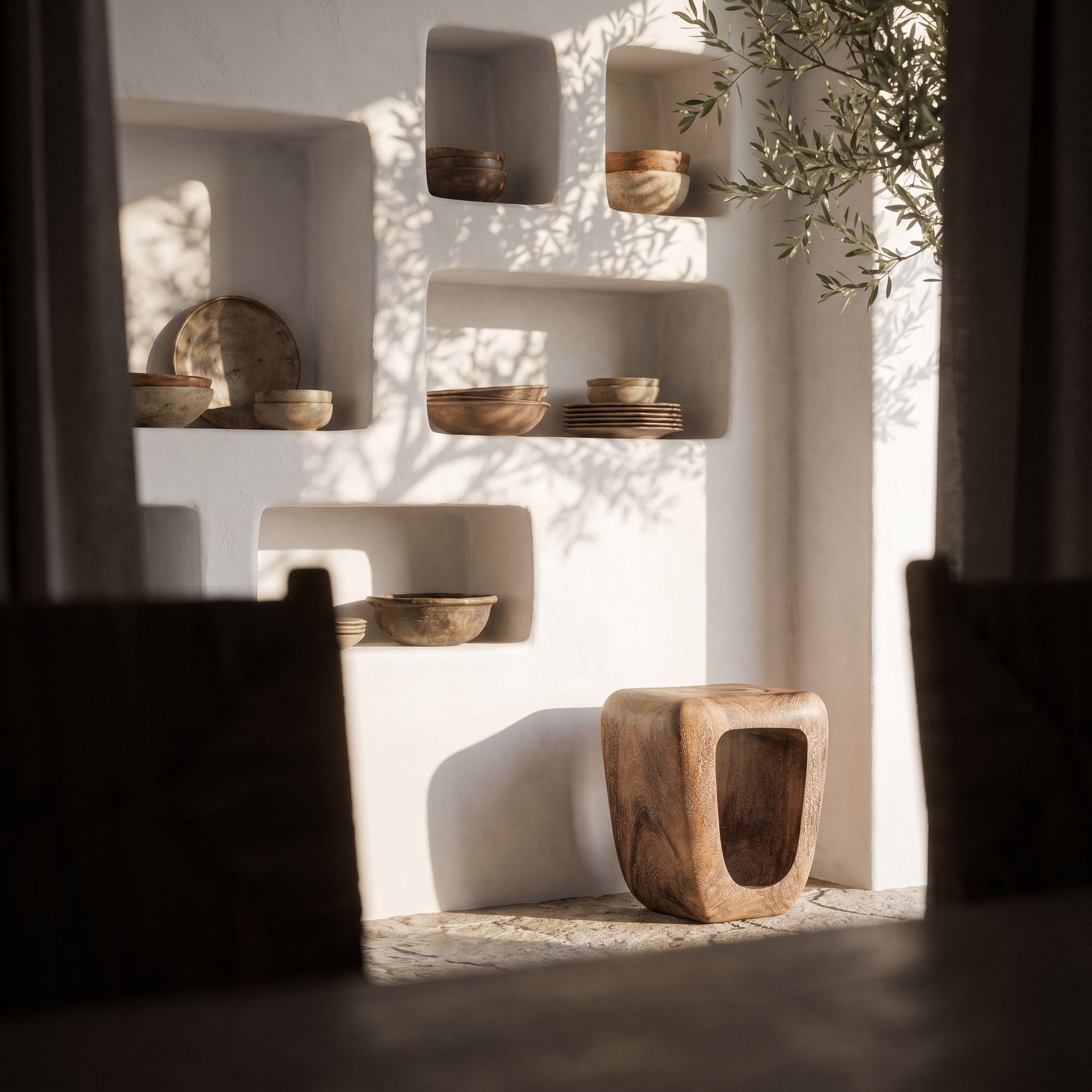 A rustic wooden stool with a hollow center sits on a stone floor in front of a white stucco wall with recessed shelves holding bowls and plates. Sunlight casts leafy shadows across the wall, with dark curtains framing the scene.