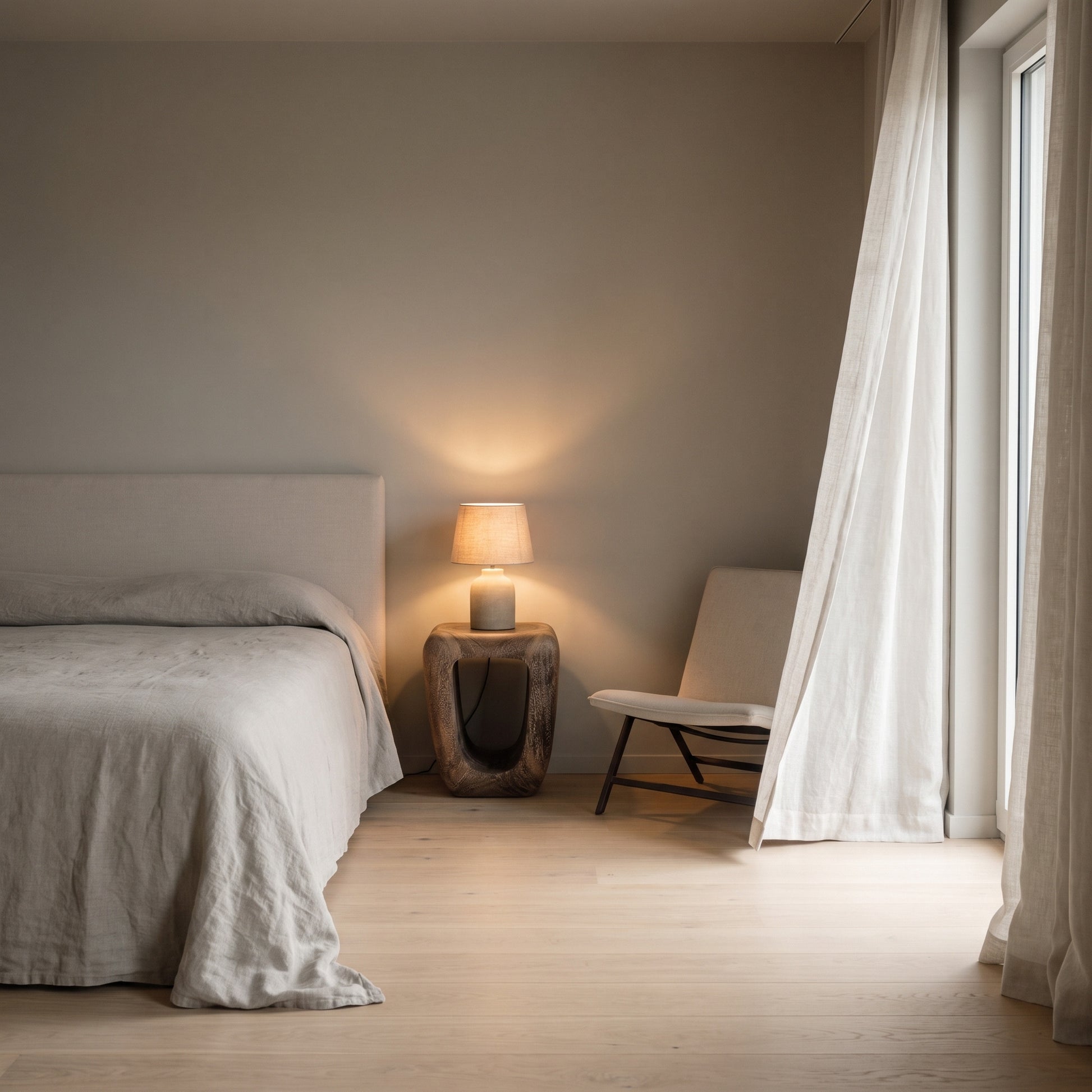 A minimalist bedroom scene with a bed dressed in light neutral bedding, a small wooden side table holding a lit lamp, a white chair, and sheer curtains by a bright window.