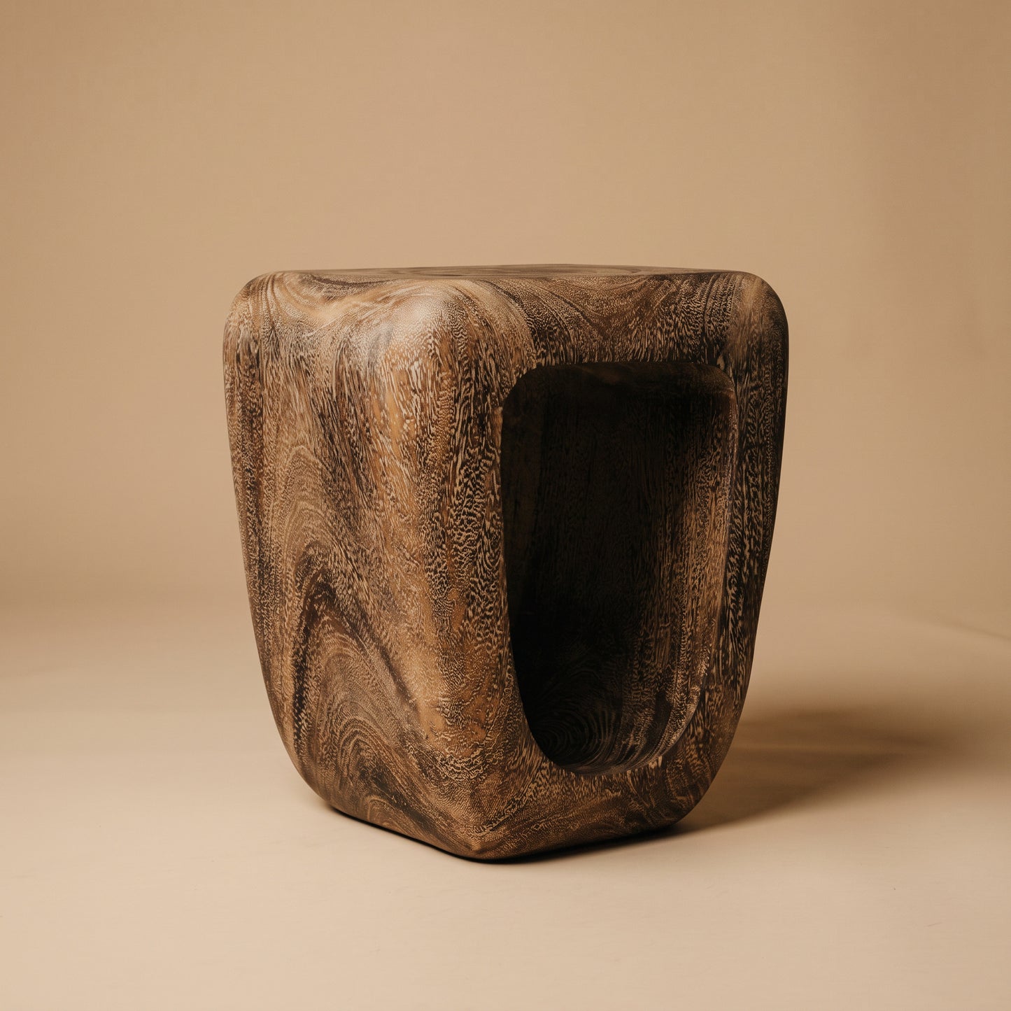 A sculptural wooden stool with a hollow center, featuring a rich dark brown wood grain pattern, set against a solid beige background.