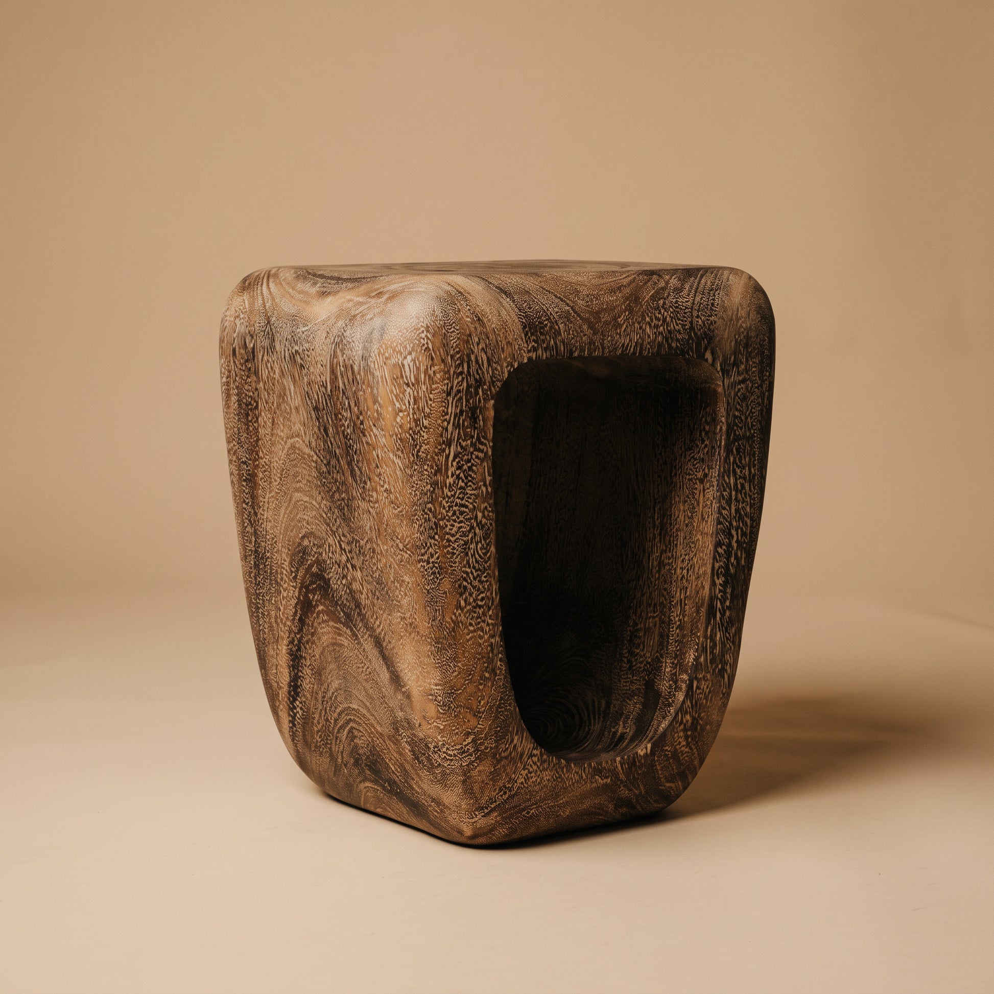 A sculptural wooden stool with a hollow center, featuring a rich dark brown wood grain pattern, set against a solid beige background.
