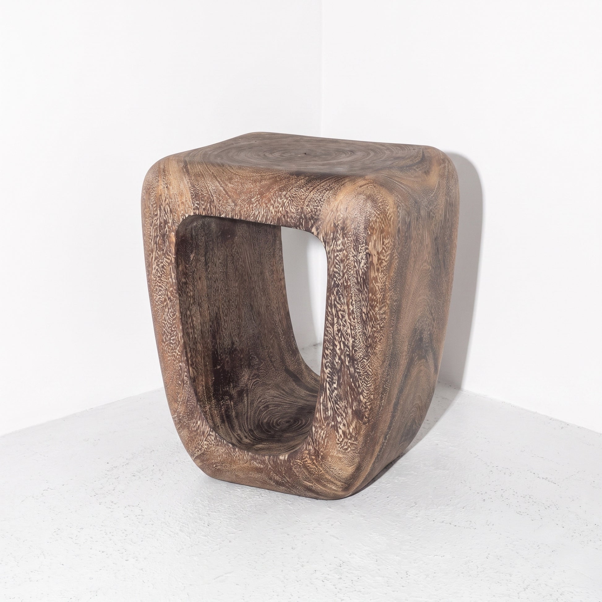 A rustic wooden side table or stool with a thick, hollow rectangular center, made from a natural dark brown wood with prominent grain patterns, photographed in a minimalist white corner setting.