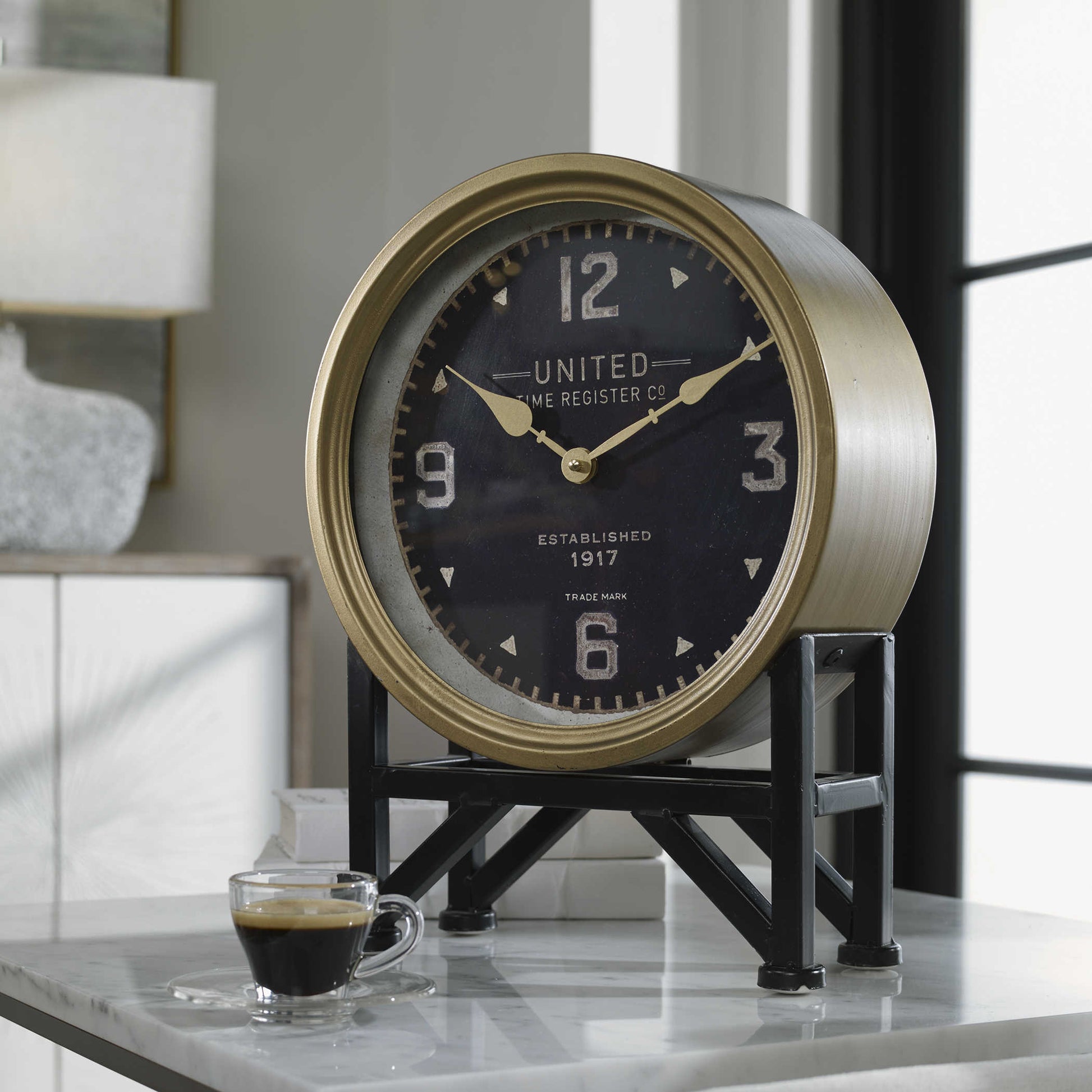 A brass-finished nautical clock with dark black accents, displaying a black clock face with white numerals and hands, placed on a glass surface possibly with a coastal decor setting.