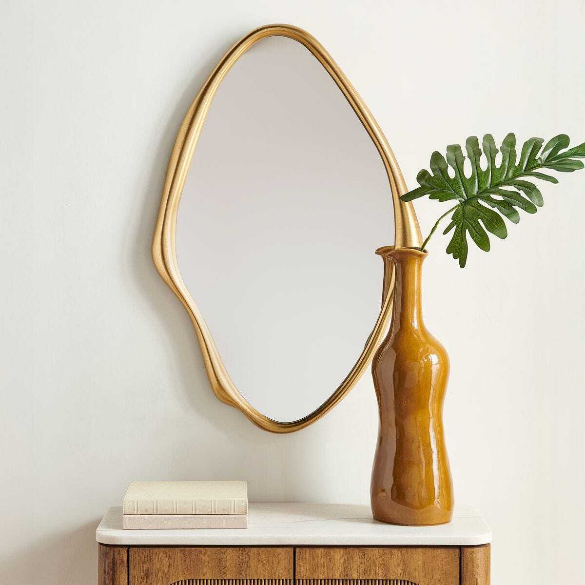 An organic-shaped wall mirror with a gold frame hangs above a wooden console table. A glossy amber-brown ceramic vase with a green monstera leaf sits on the table next to a small stack of books.
