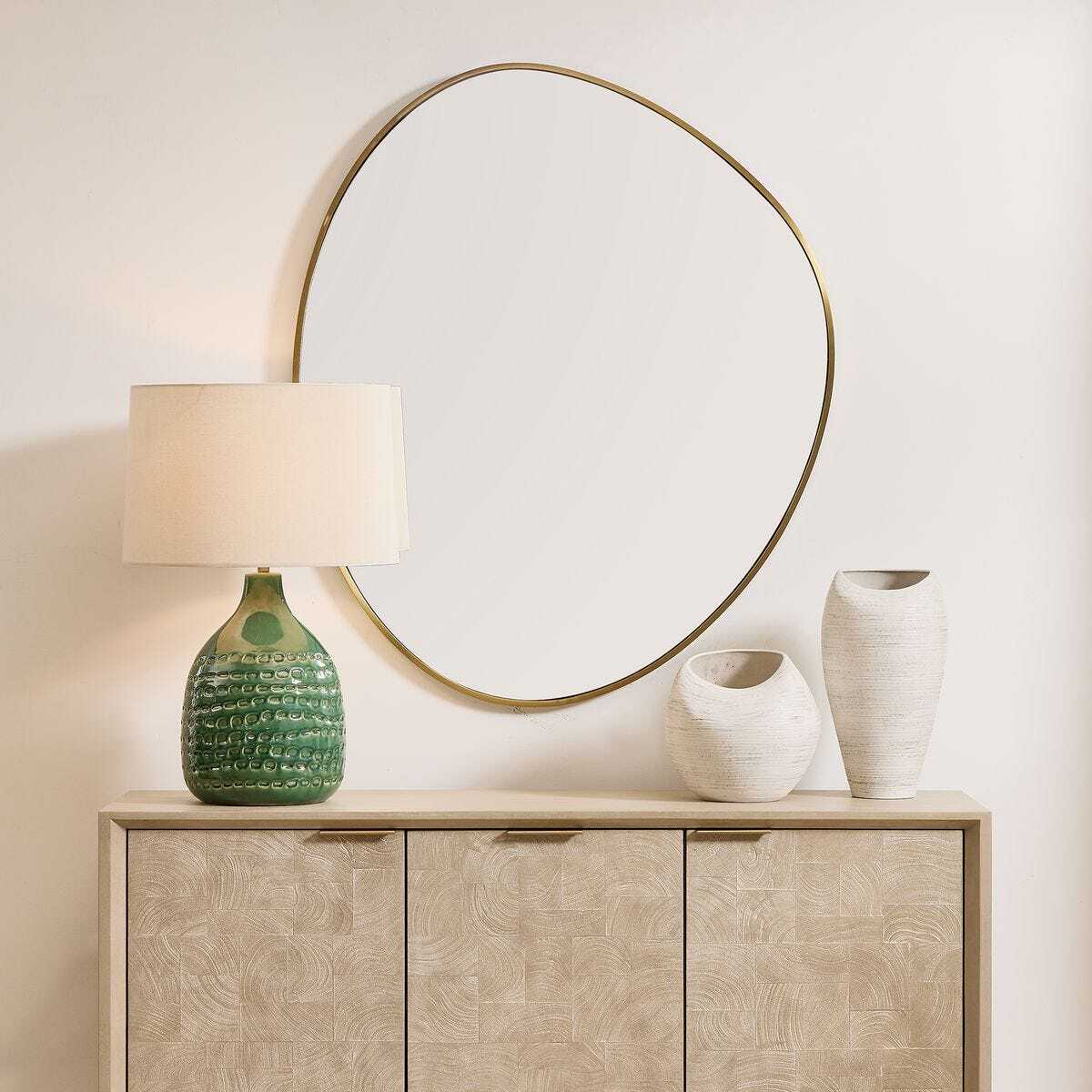 Round mirror with gold frame above a wooden cabinet with decorative vases and a lamp.
