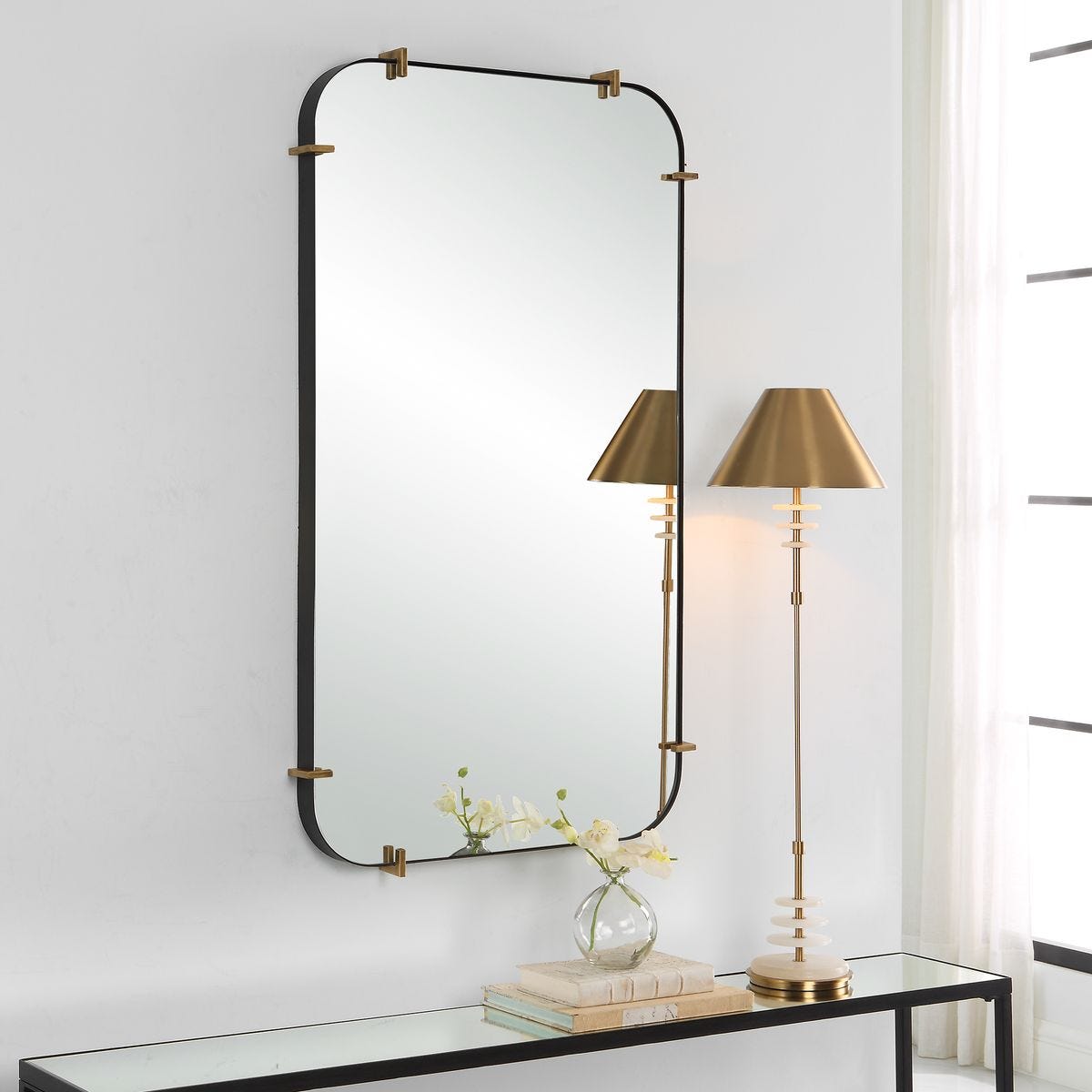 Decorative mirror with gold frame on a white wall above a glass table with books and a vase.