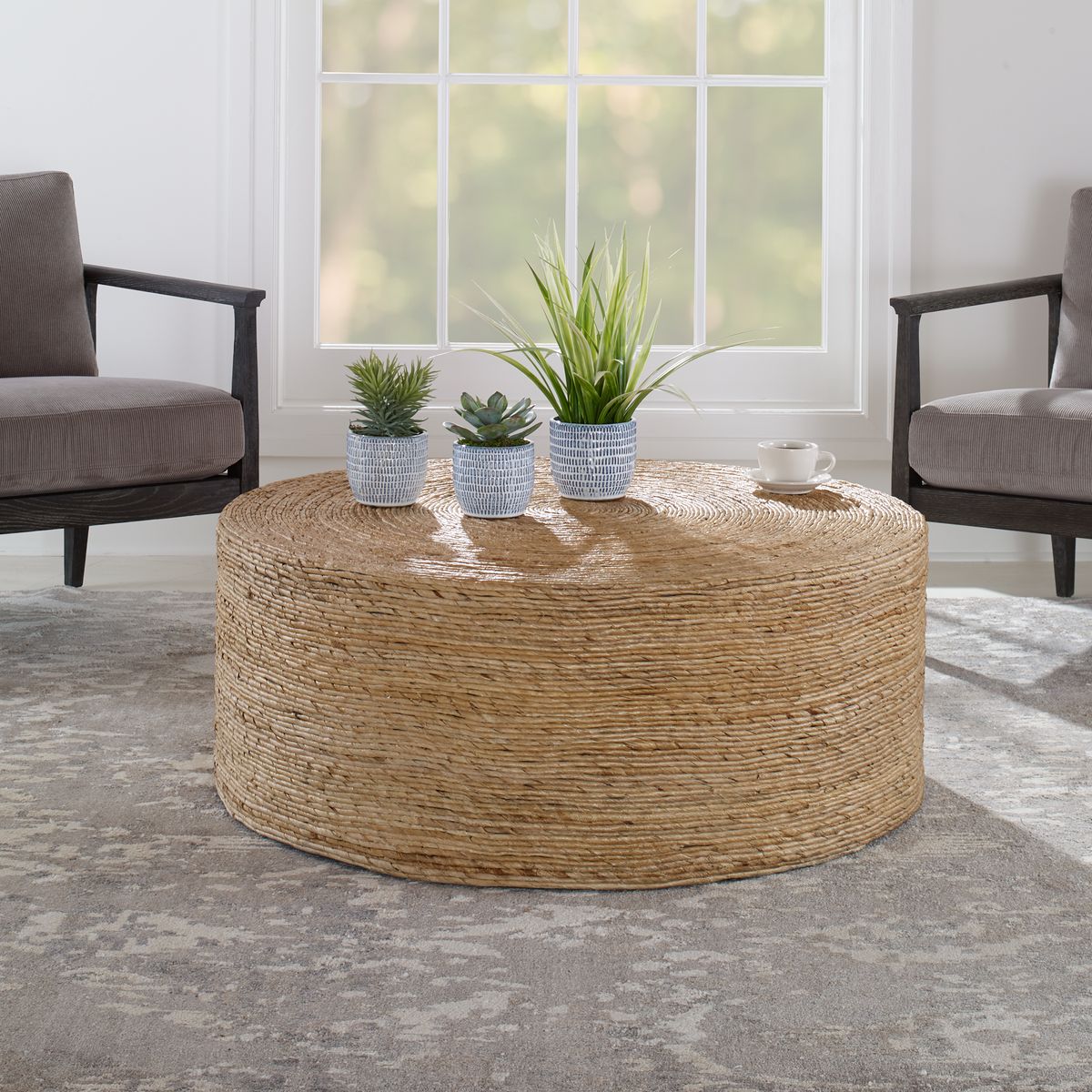 A round coastal coffee table made of woven banana leaf, placed on a grey area rug in a room with two gray chairs and several potted plants on top of the table.