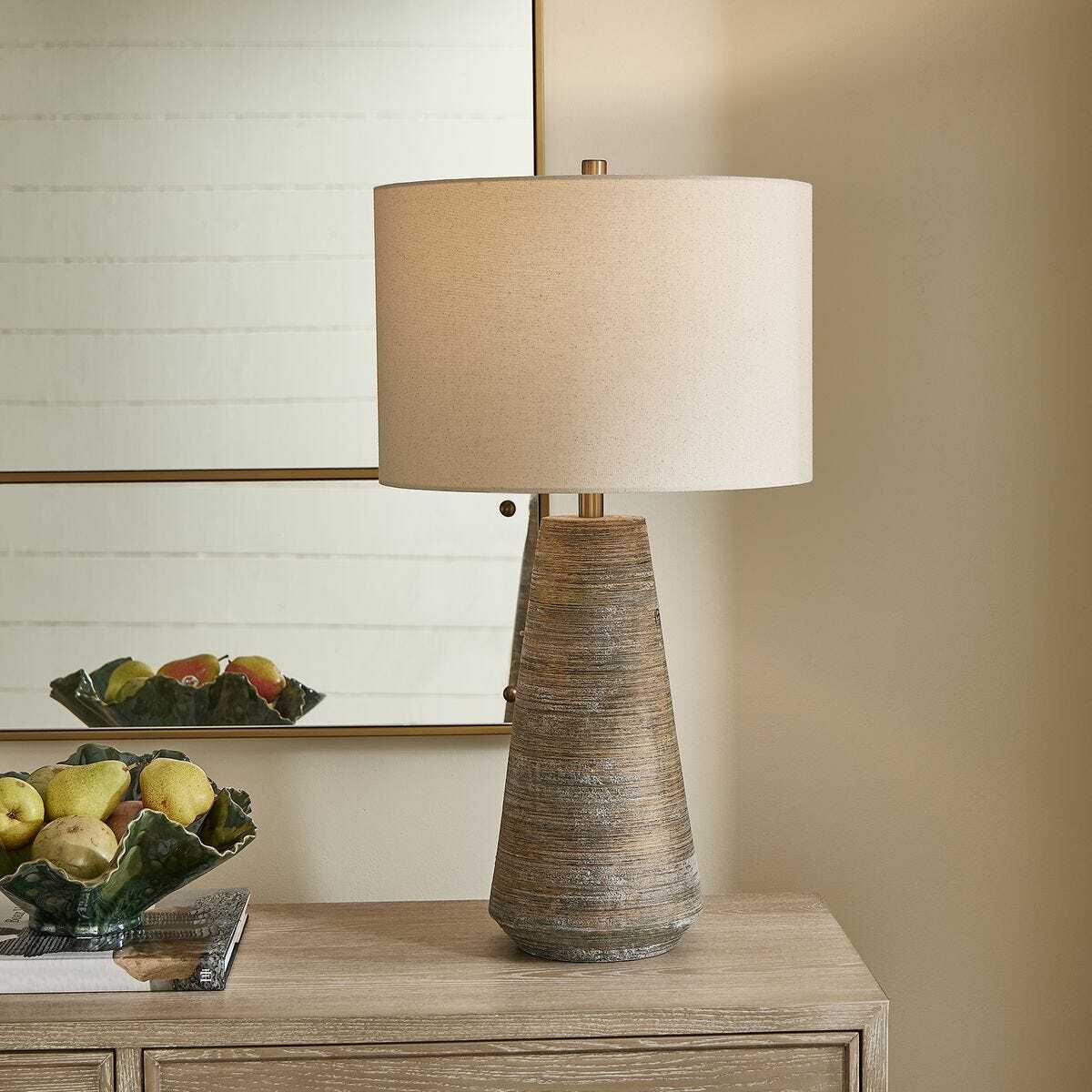 Decorative lamp with a textured base on a wooden surface, with a fruit bowl and abstract painting in the background.