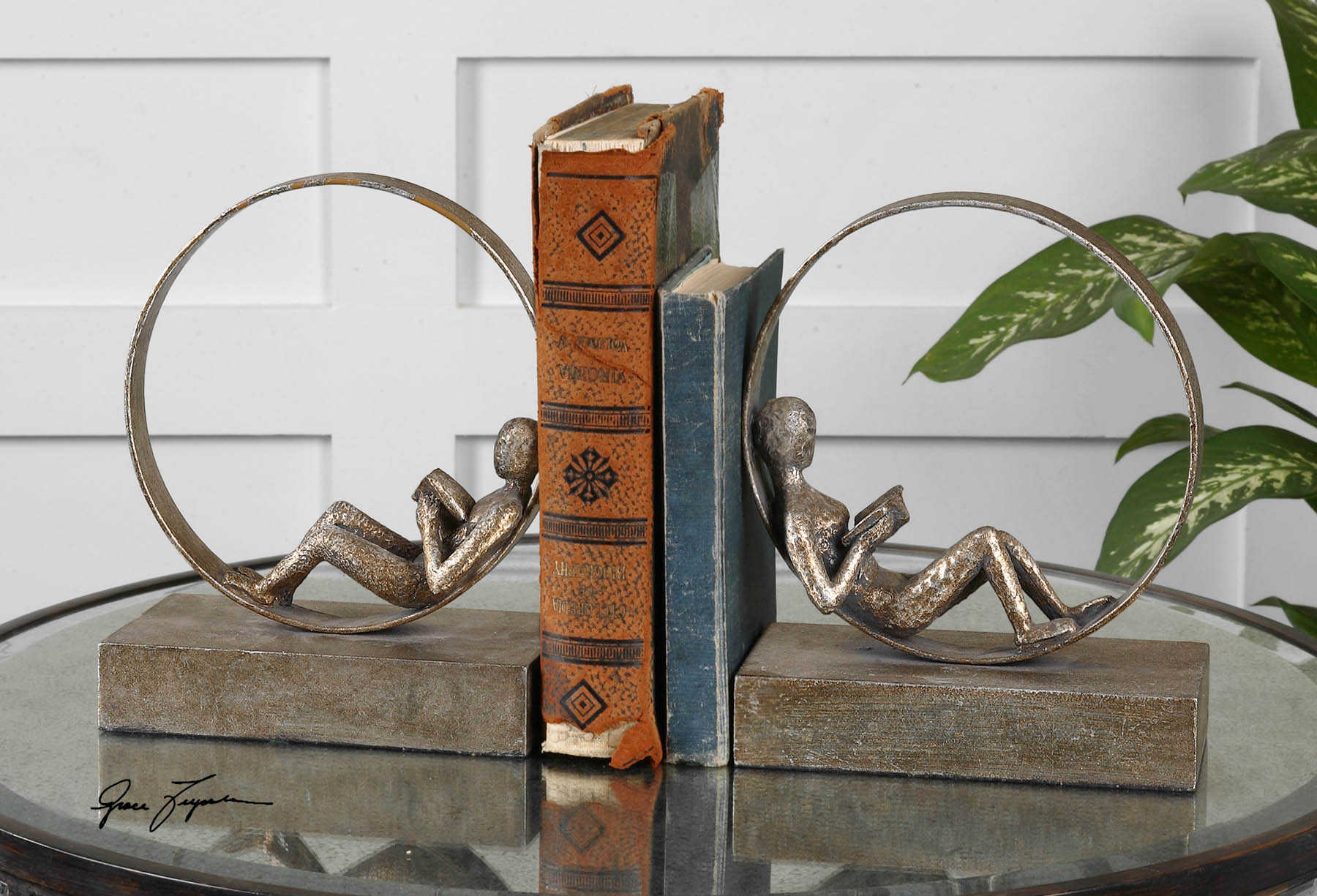 A pair of silver bookends designed as stylized seated figures holding bookends, placed on a glass surface with a book positioned between them.