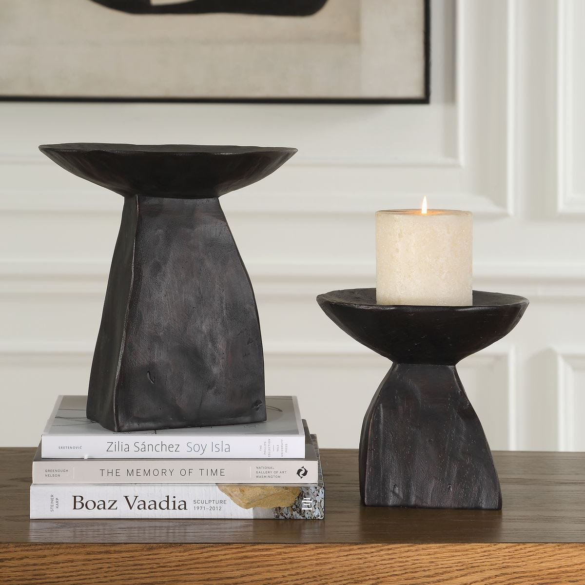 Two pedestal bowls with a matte dark brown finish, one small and one large, displayed on a surface with a candle on the smaller one and books stacked under the larger one.