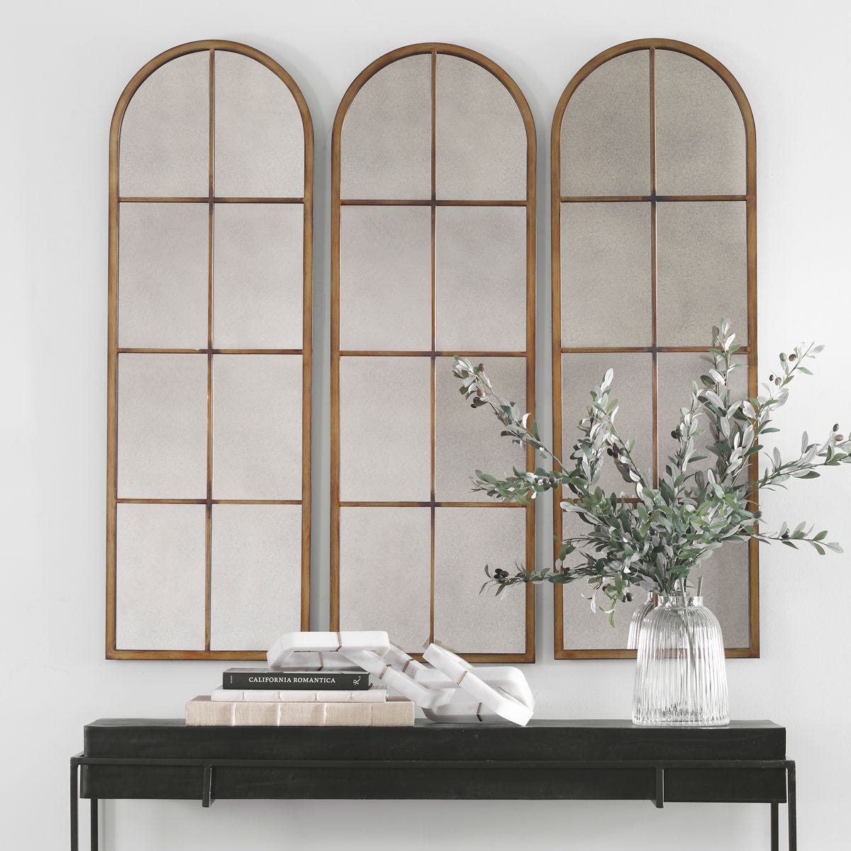 Three arched mirrors with a vase of flowers on a console table against a white wall.
