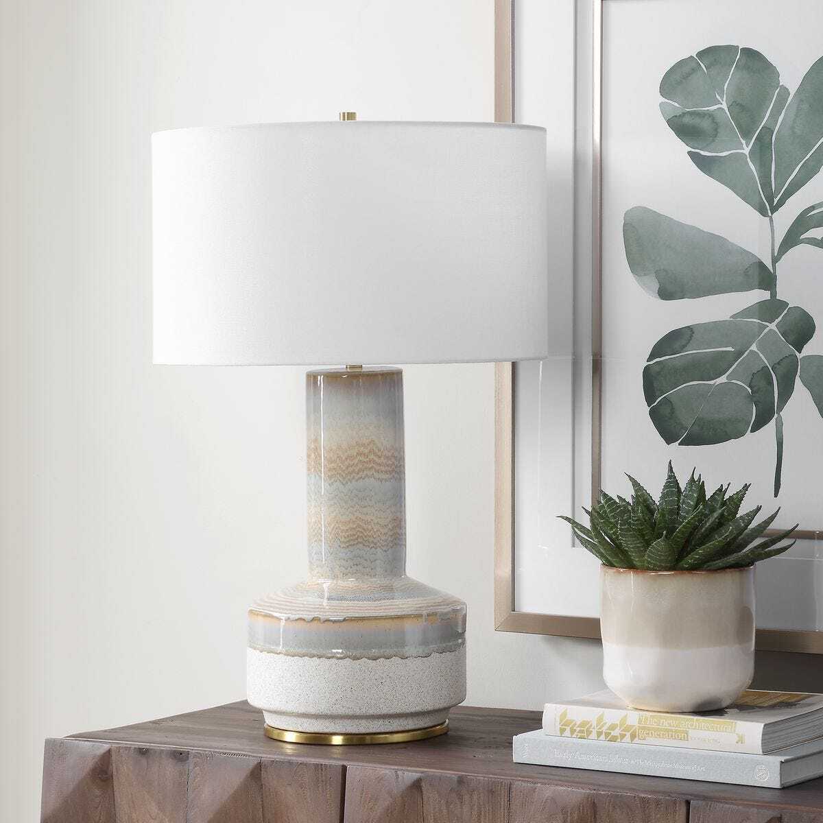 Table lamp with a decorative base on a wooden surface next to a plant and books.