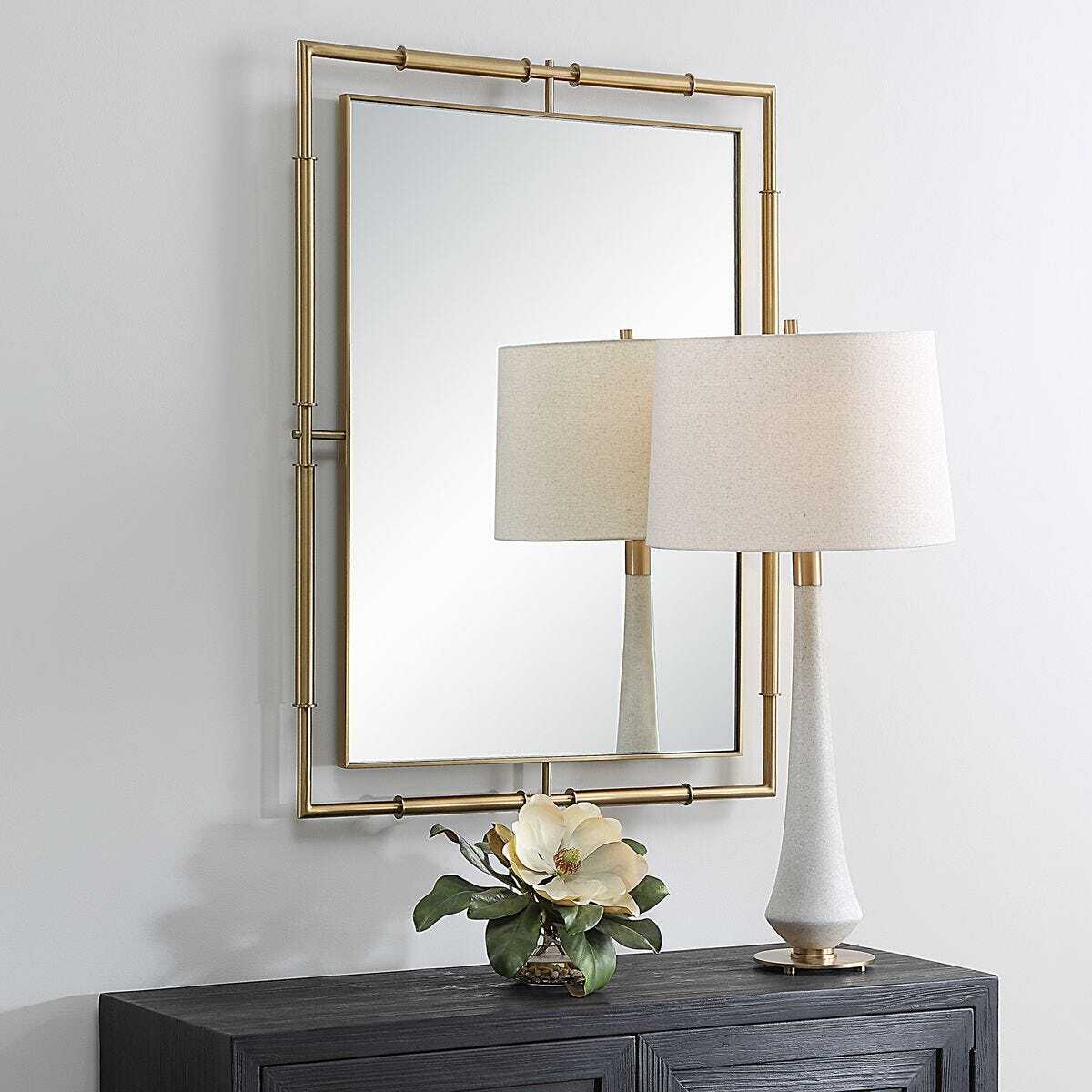 A rectangular wall mirror with a gold-toned metal frame hangs above a dark wood console table. On the table sits a white ceramic table lamp with a beige fabric drum shade and a small vase with a large white flower.