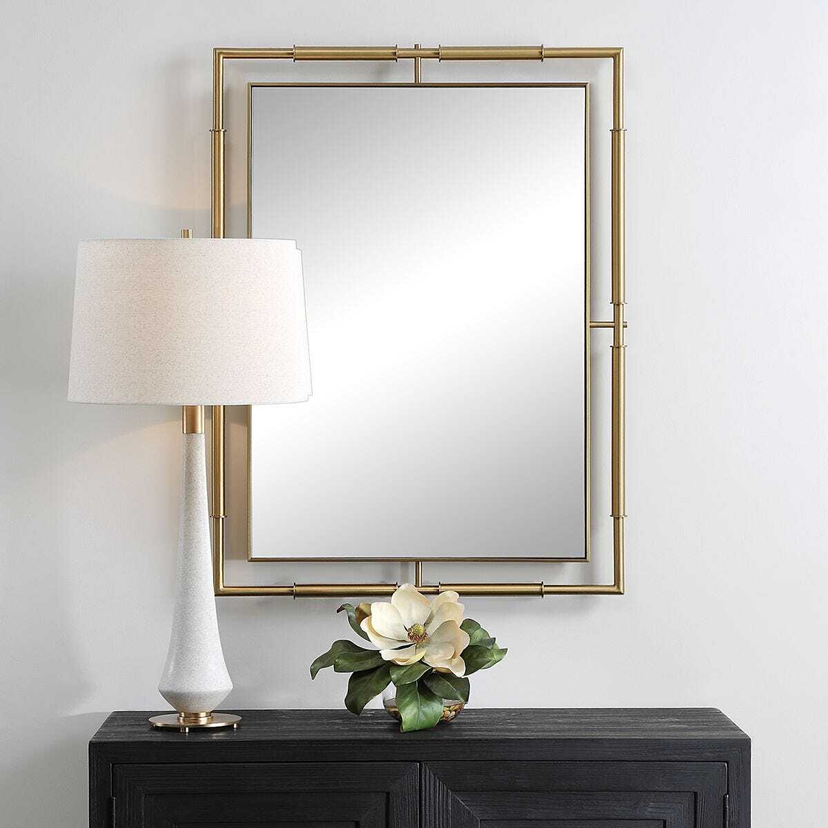 Decorative mirror with gold frame above a dark wooden console table with a lamp and floral arrangement.