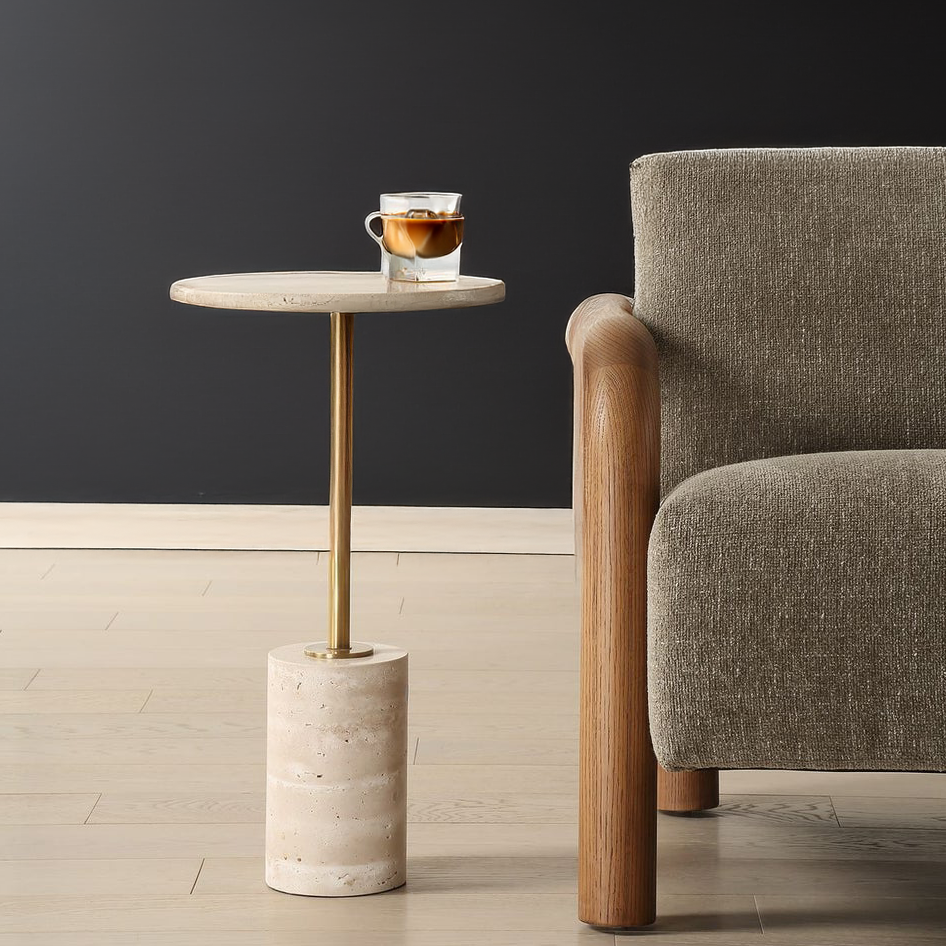 Travertine side table with glass of coffee and a gold stem next to a chair in a black living room.