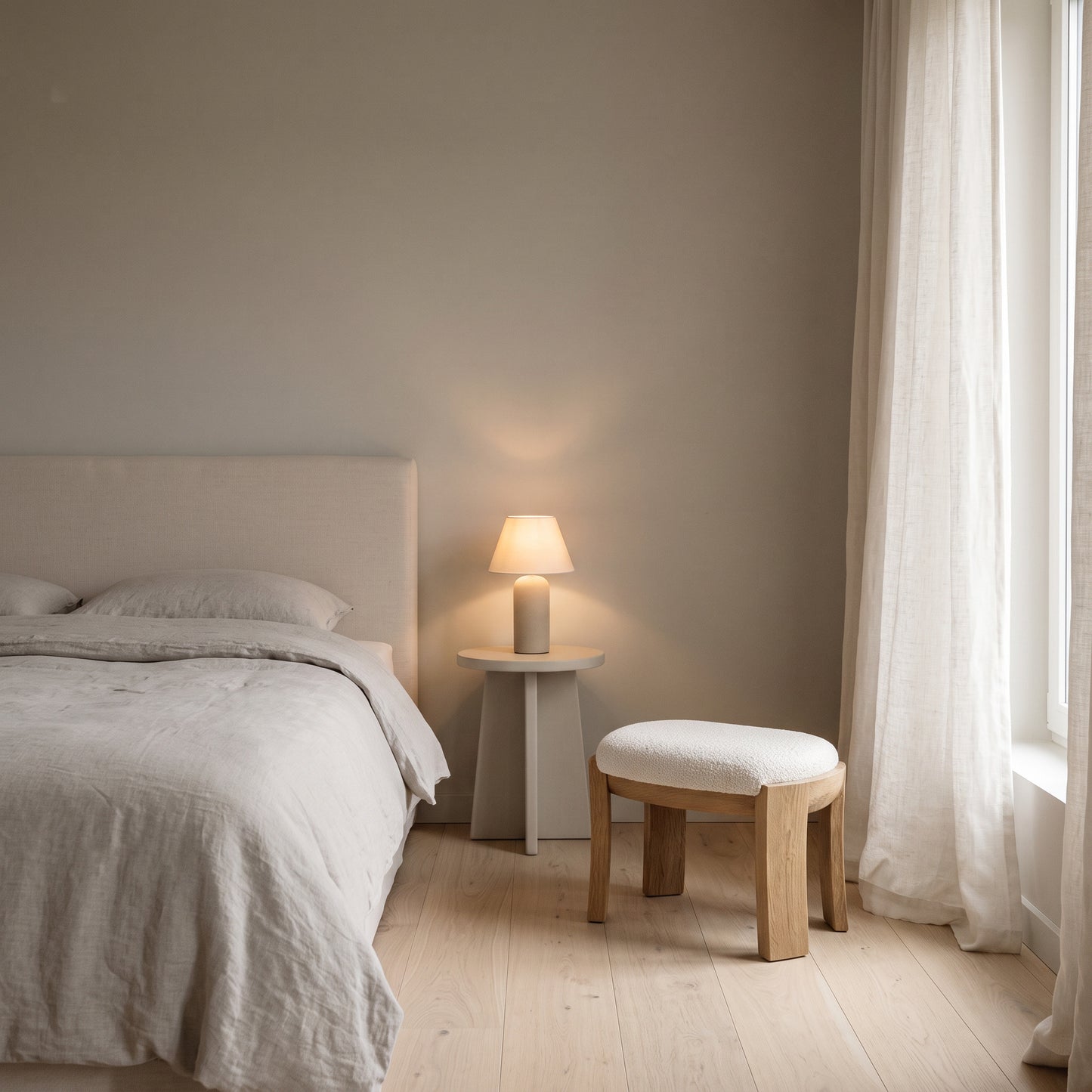 A softly lit bedroom corner with a bed in white linens, a small side table with a warm lamp, a wooden stool with a white cushion, and sheer white curtains against a neutral beige wall.
