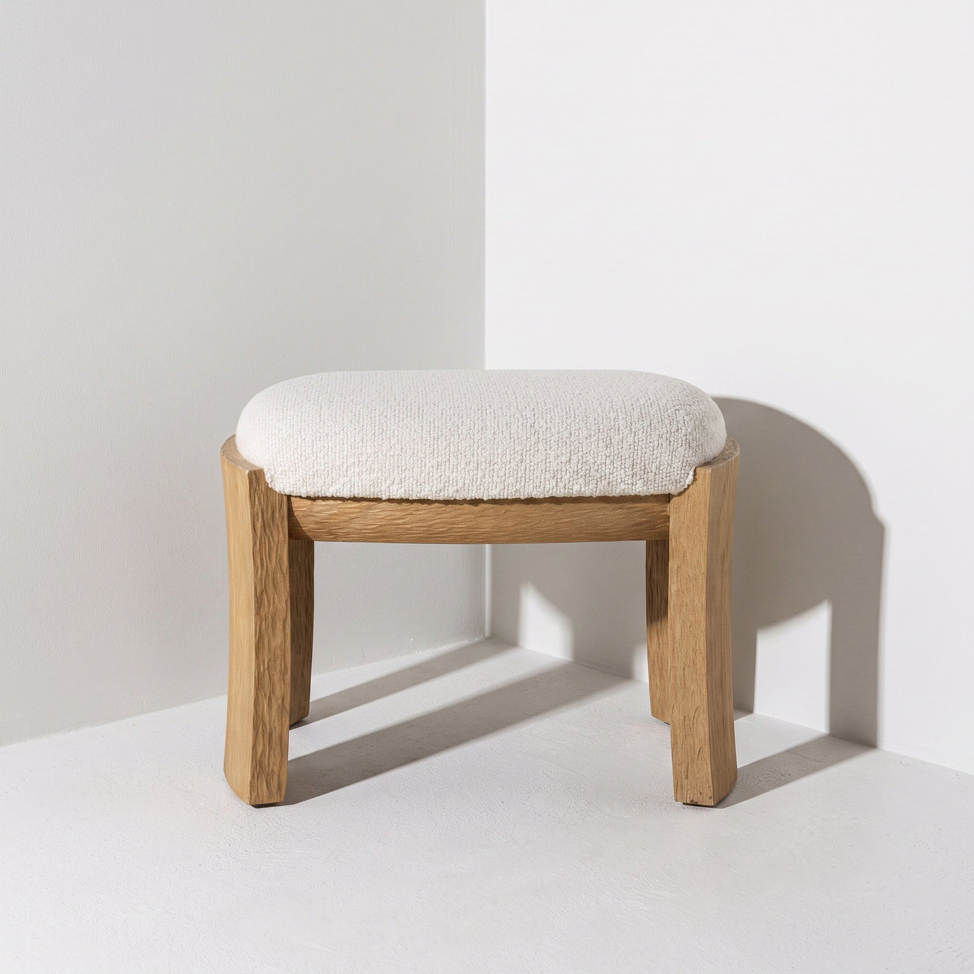 A small wooden stool with a thick, textured off-white bouclé seat cushion, photographed in a minimalist white studio corner with a soft shadow.