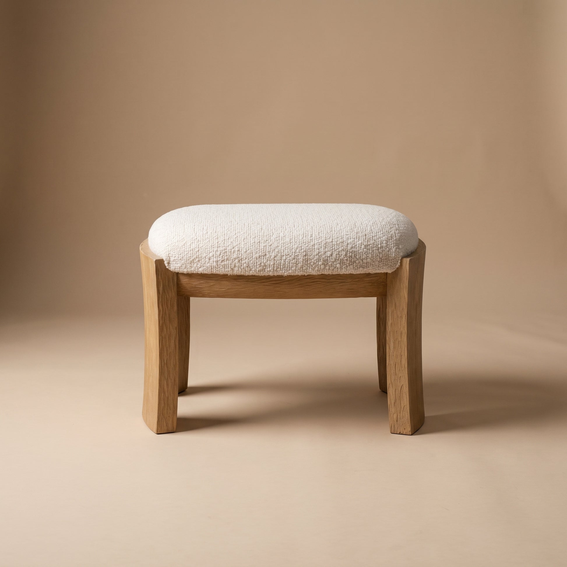A small wooden stool with a thick, textured off-white bouclé seat cushion, photographed against a seamless warm beige studio background.