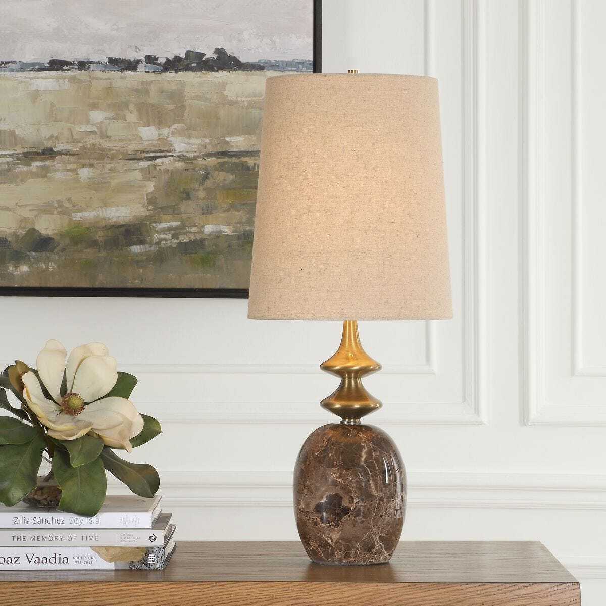 Decorative lamp with a textured base on a wooden surface, with books and a floral arrangement in the background.