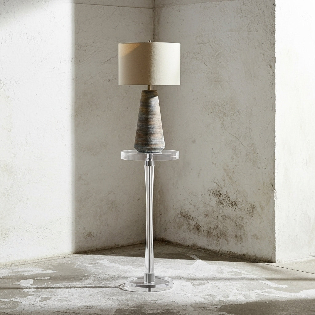 Table lamp with a textured base and beige shade on a clear stand against a textured wall.