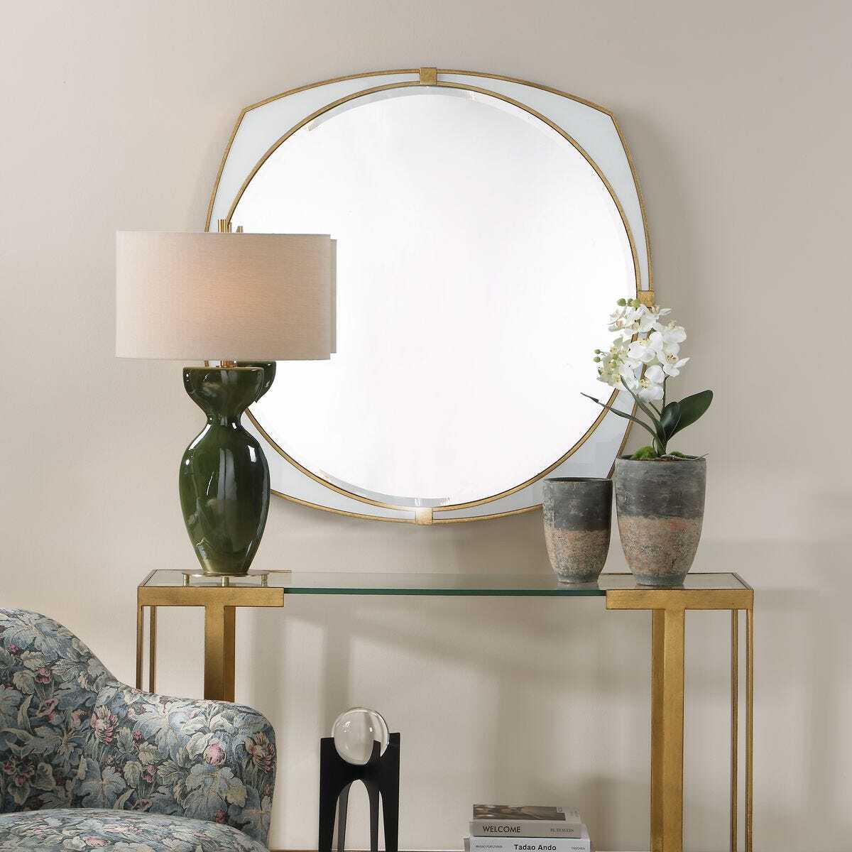 An octagonal wall mirror with a gold frame hangs above a glass-top console table in a styled interior. A dark green table lamp, two textured vases, and a white orchid are placed on the table, with part of a floral armchair visible at the lower left.