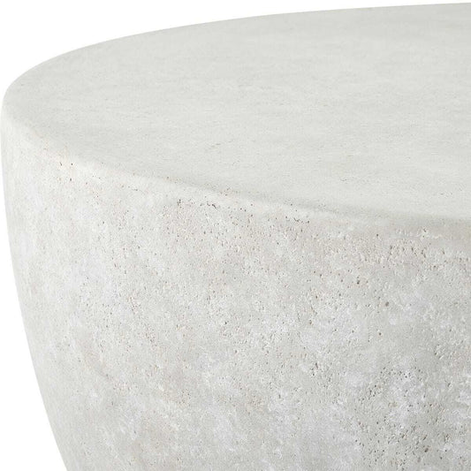 A round, low-profile coffee table made of textured concrete or stone in a light warm gray/beige tone, positioned on a stone patio between two modern outdoor sofas.