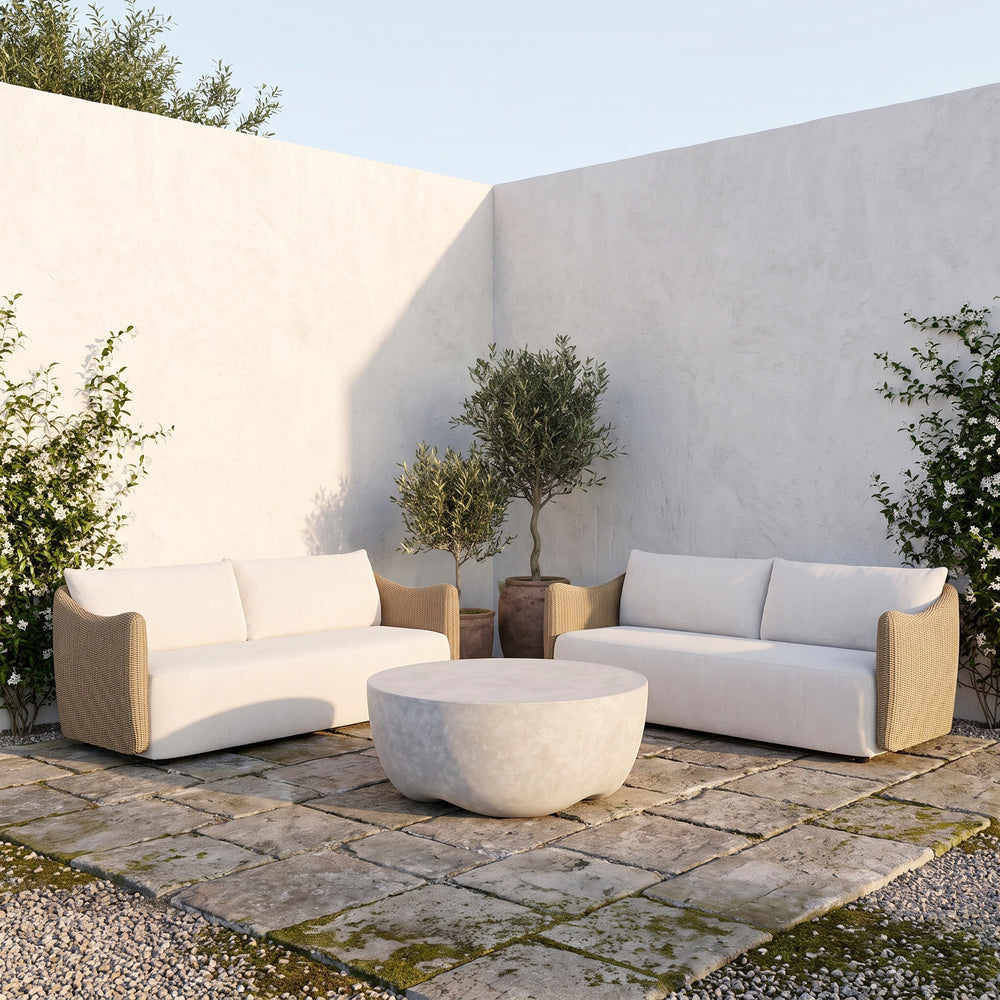 Modern outdoor patio furniture set featuring two beige armchairs with white cushions and a matching round stone coffee table, arranged on a stone patio against a white stucco wall.