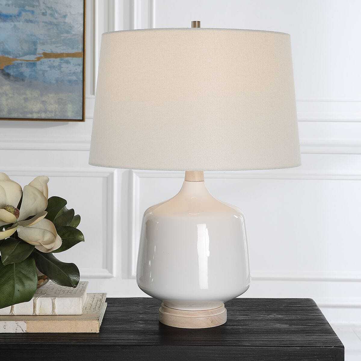 Table lamp with beige shade on a wooden surface, with books and a plant in the background.