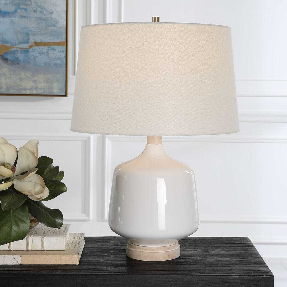 Table lamp with beige shade on a wooden surface, with books and a plant in the background.