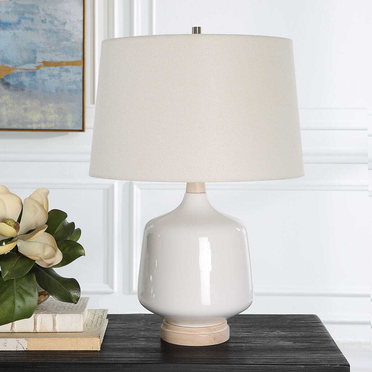 White table lamp with beige shade on a wooden surface, with books and a plant in the background.