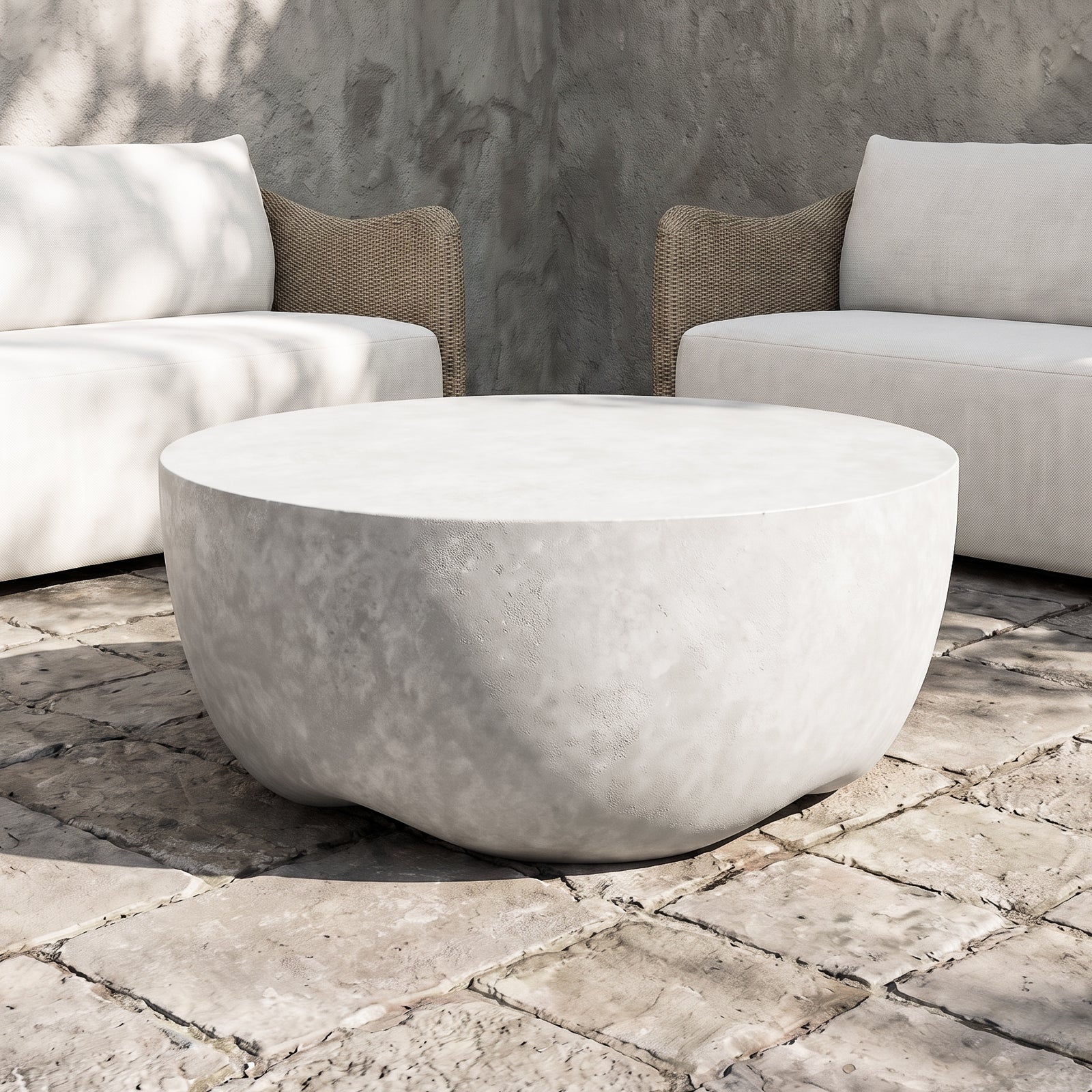 A round, low-profile coffee table made of textured concrete or stone in a light warm gray/beige tone, positioned on a stone patio between two modern outdoor sofas.