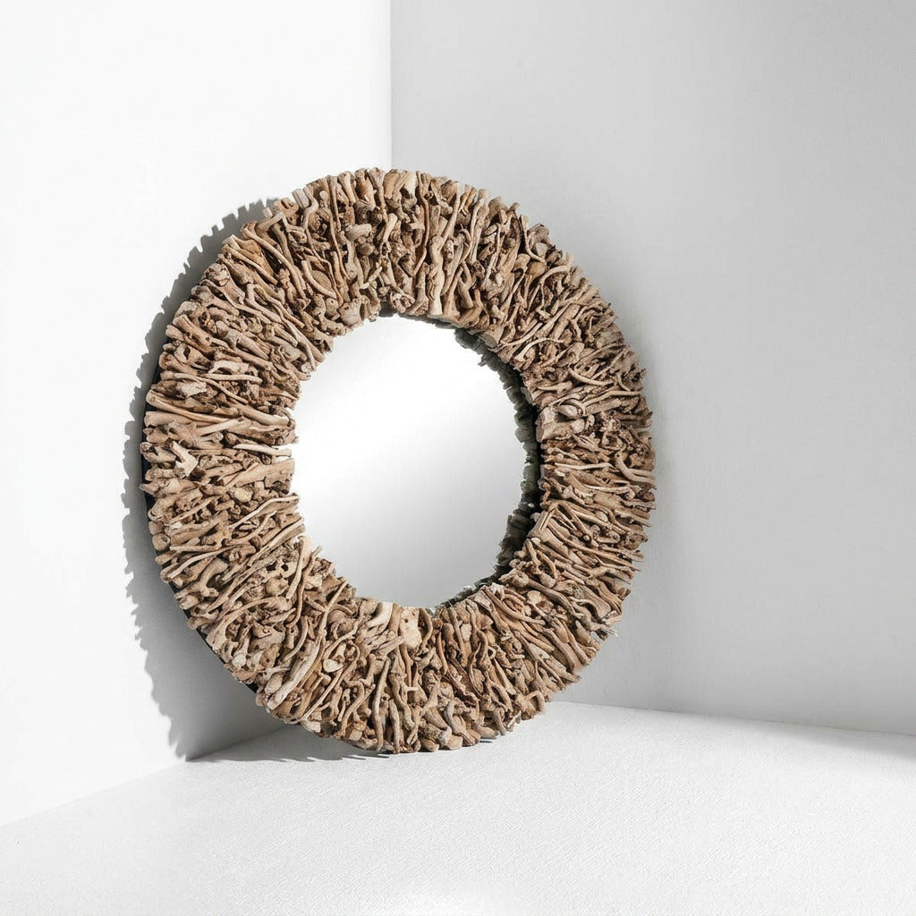 An oval decorative mirror with a thick, textured natural wood or driftwood-style frame, leaning against a white wall in a minimalist corner setting.