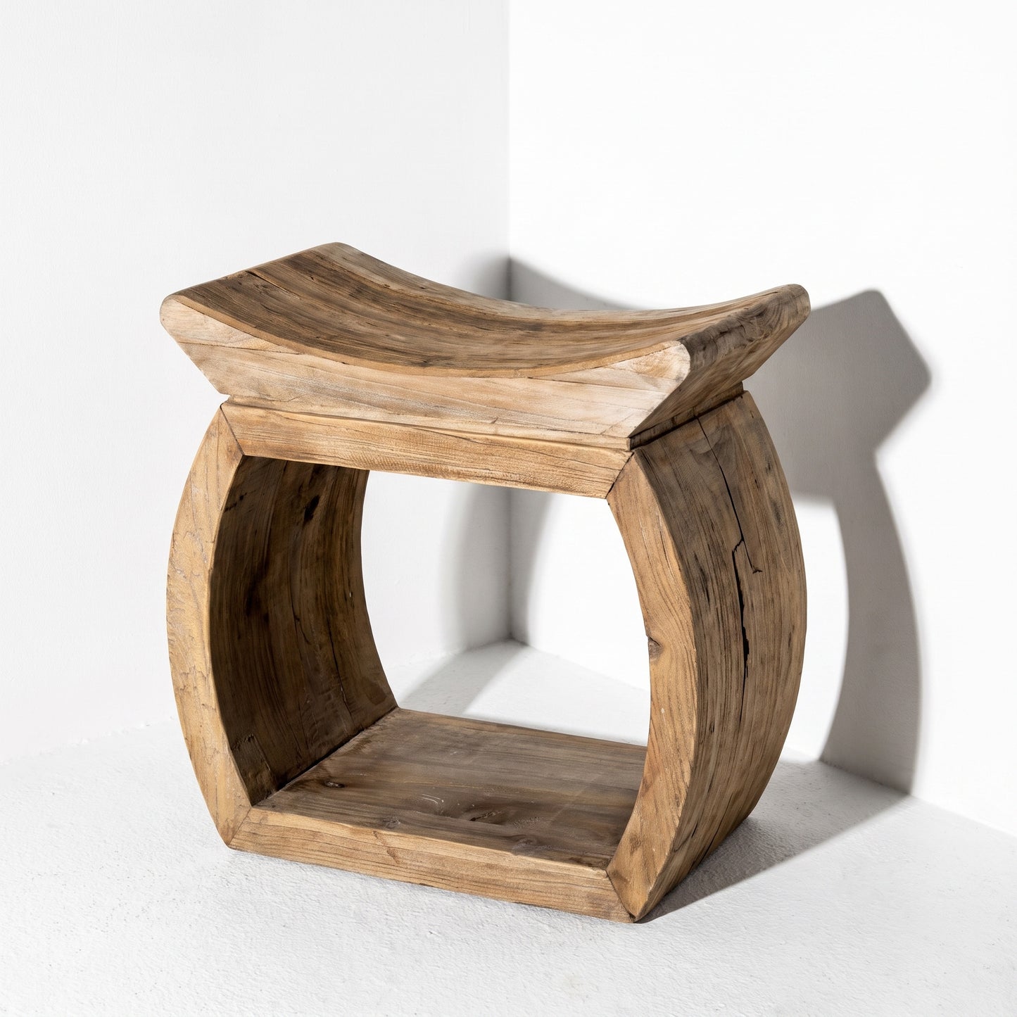 A rustic wooden stool with a curved, open-frame design and a gently curved seat, shown in a minimalist white studio corner with a soft shadow.
