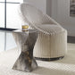 Silver twist accent table with a glass of whiskey on top next to a decorative chair on a textured white rug. 